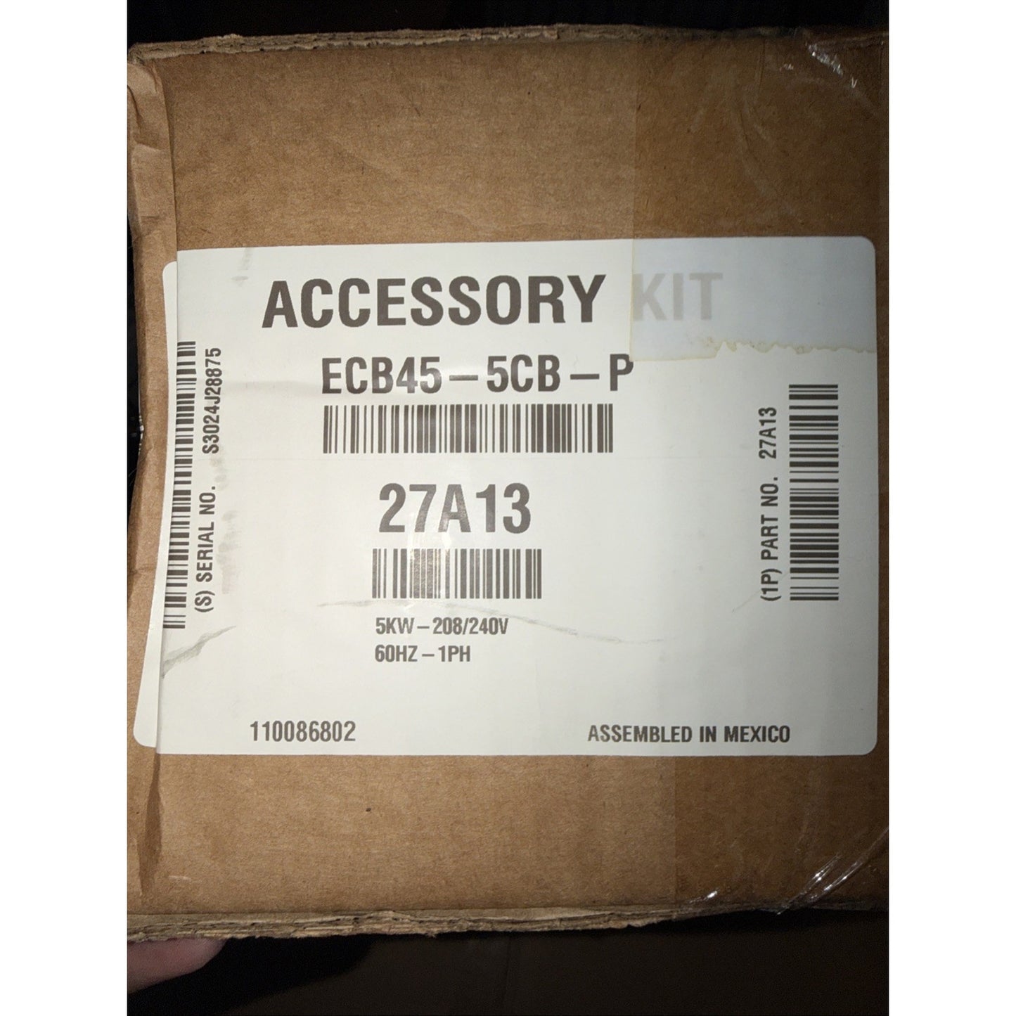 Brown cardboard box with a white label displaying an accessory kit code, barcode, electrical specifications of 5kW 208/240V 60Hz 1PH, and assembled in Mexico information.