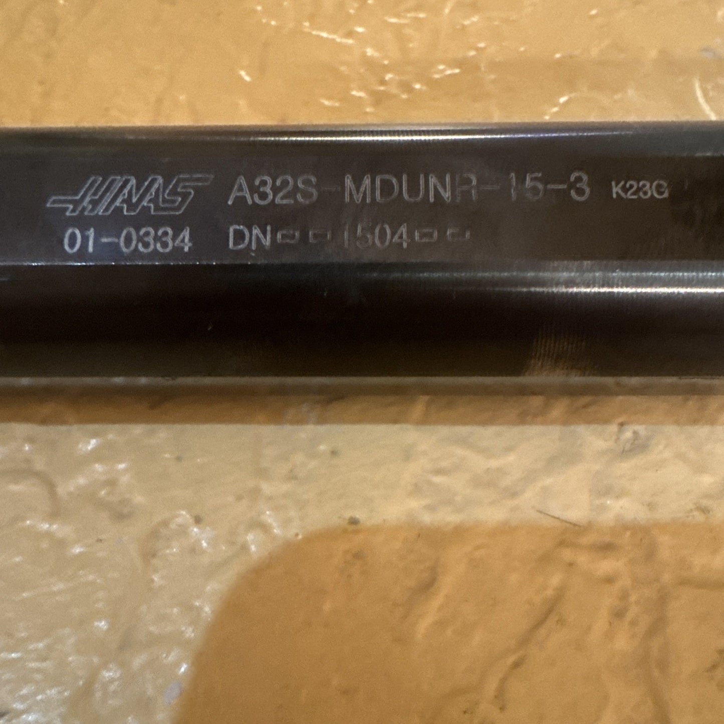 Close-up of a dark metal tool shaft engraved with the Haas logo, model code A32S-MDUNR-15-3, and additional alphanumeric markings, resting on a textured beige surface.