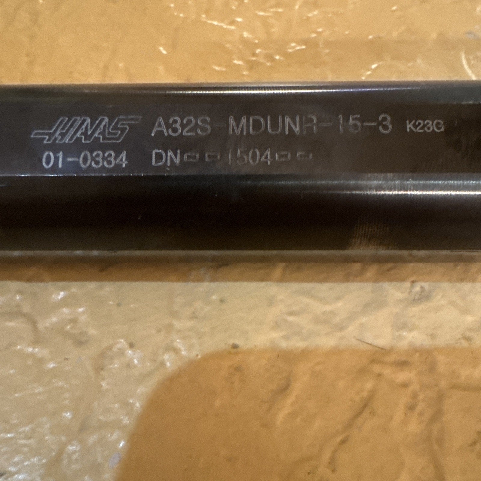Close-up of a dark metal tool shaft engraved with the Haas logo, model code A32S-MDUNR-15-3, and additional alphanumeric markings, resting on a textured beige surface.