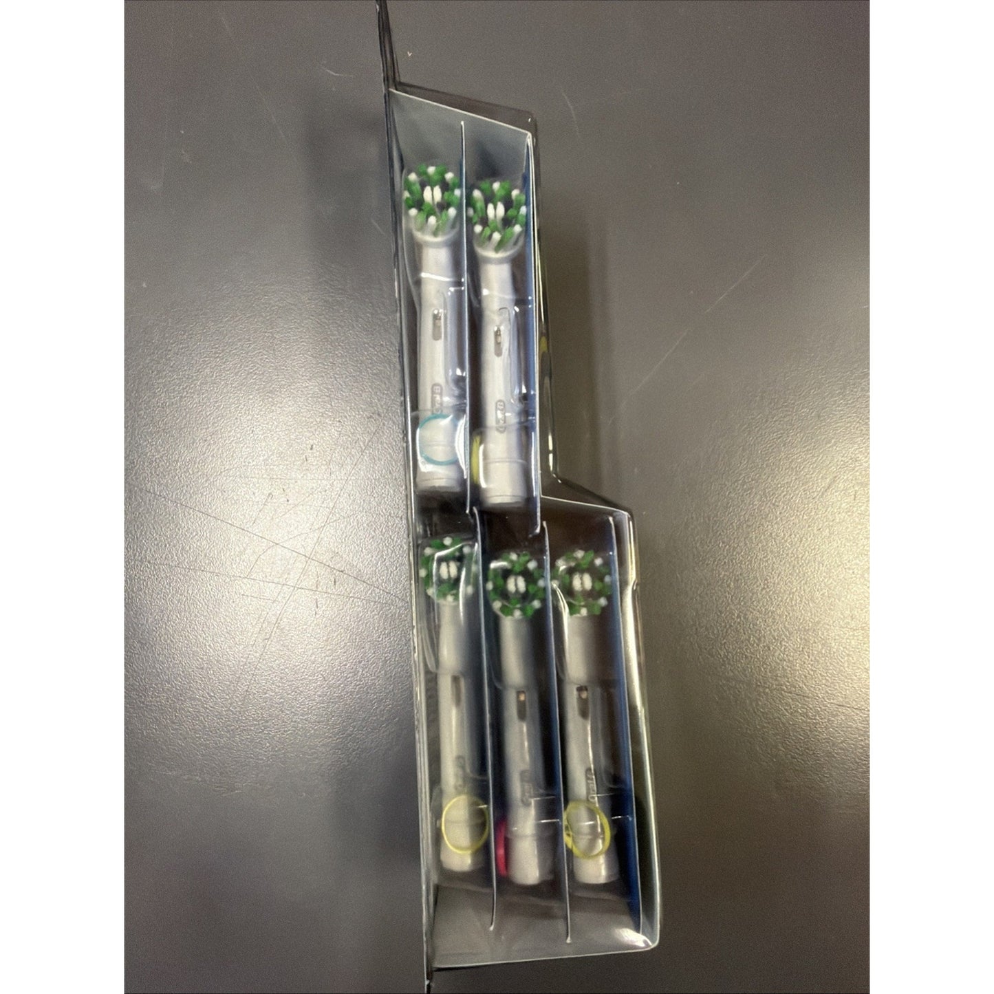 Clear plastic packaging holding five white electric toothbrush replacement heads with green and white bristles, displayed on a dark matte surface.