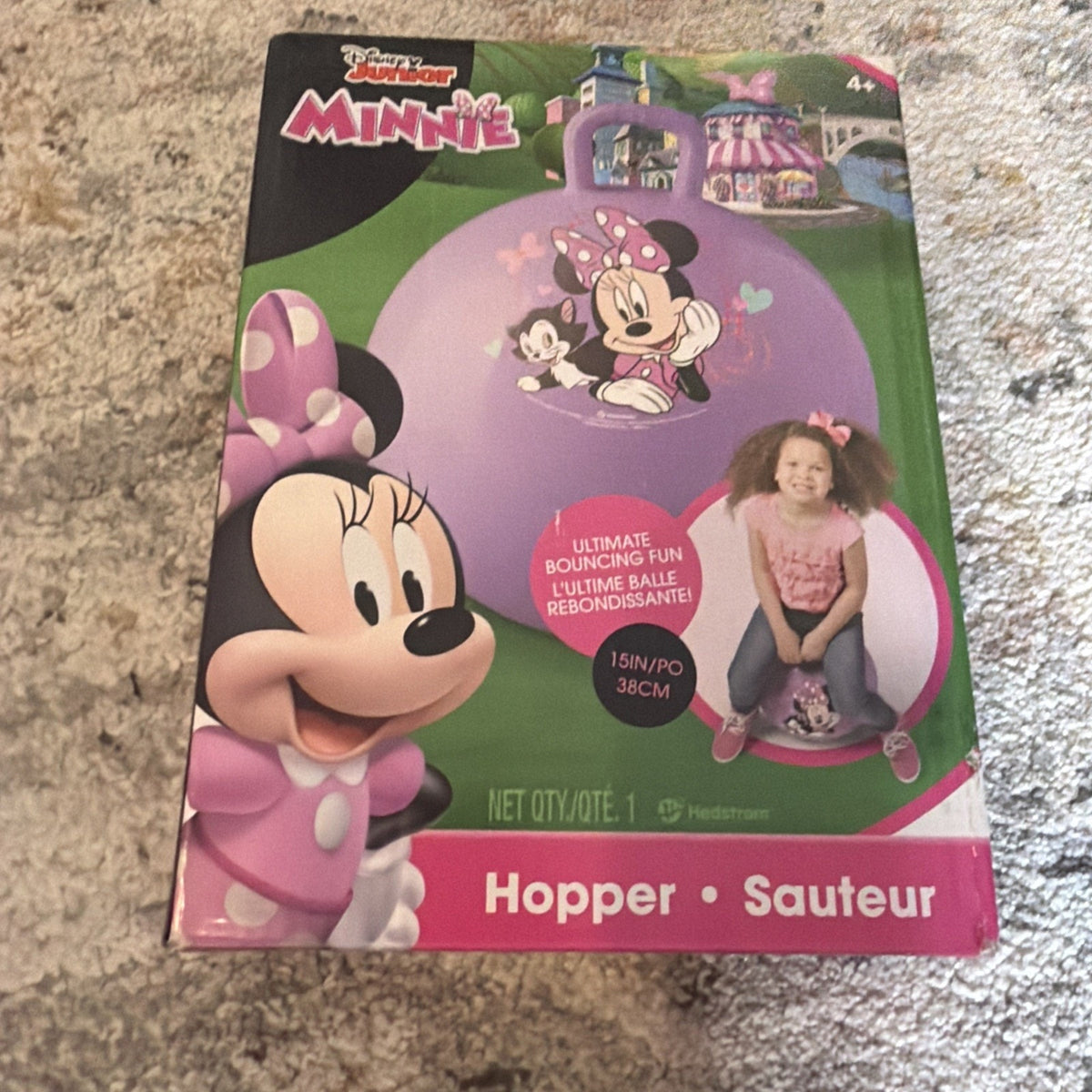 Box packaging showing a purple bouncy hopper ball featuring Minnie Mouse graphics, with a photo of a young girl sitting and bouncing on the ball, placed on a carpeted floor.