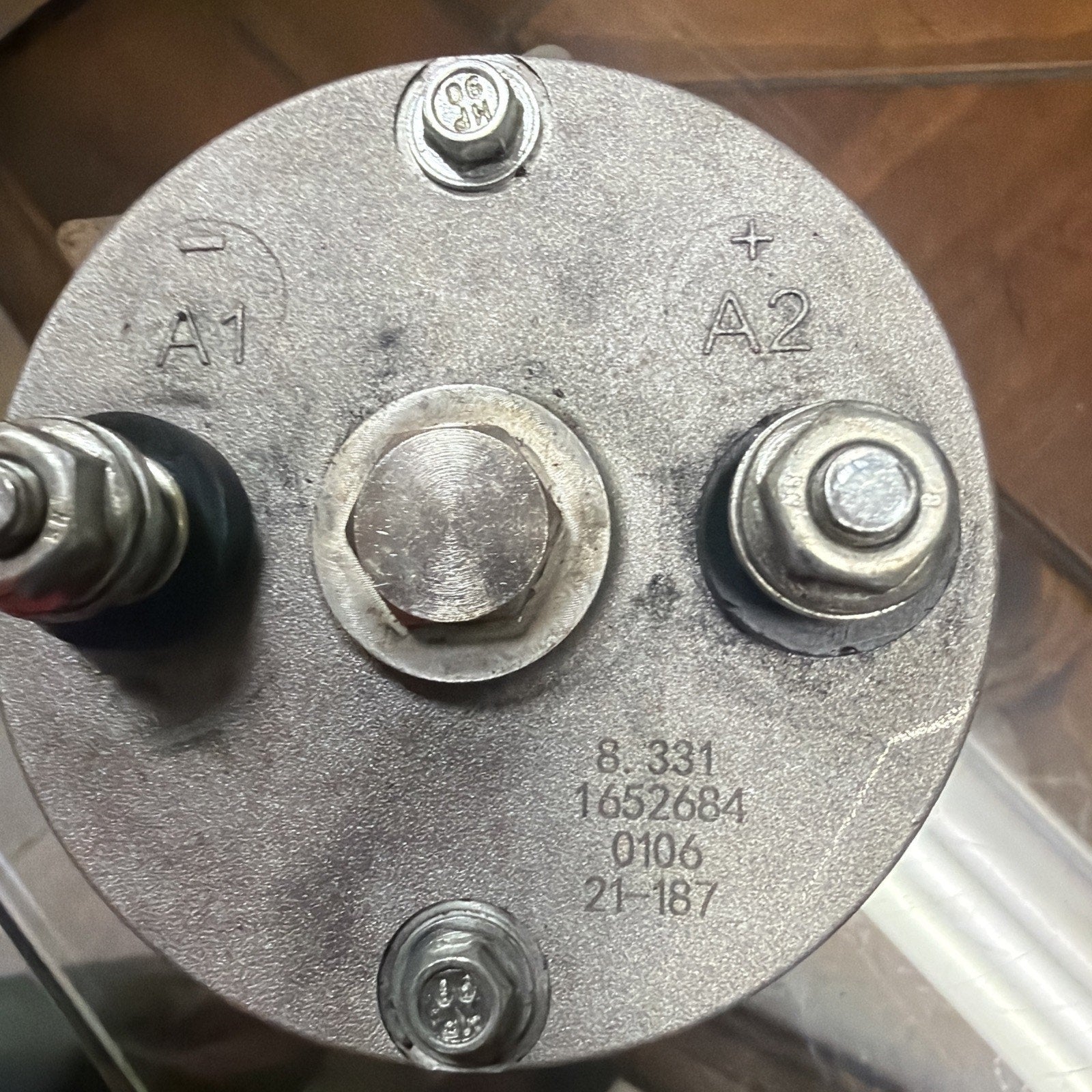 Top view of a circular metallic electrical component with two large terminals labeled A1 and A2, two smaller screws, and engraved numerical markings on its surface.