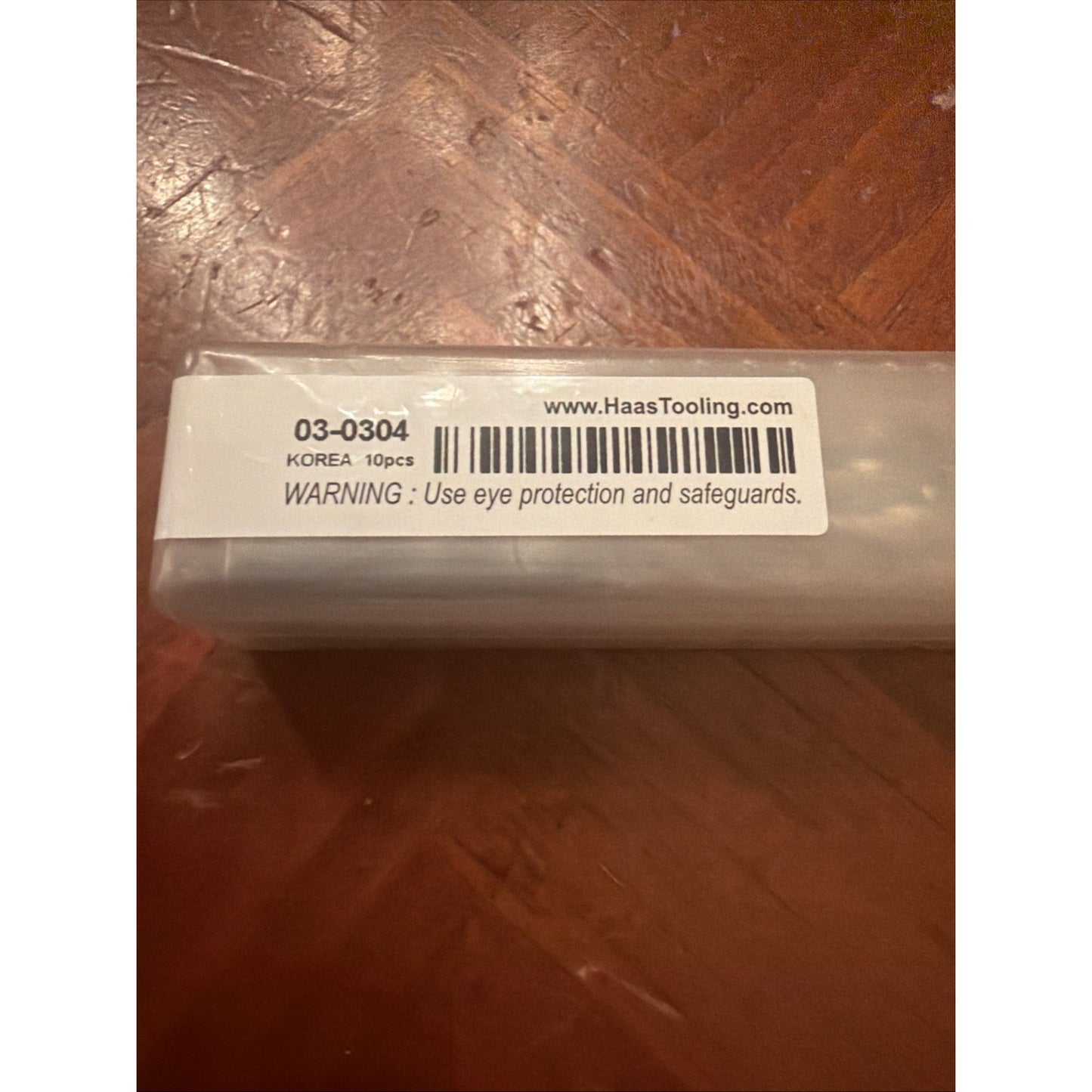 Close-up of a white label on a clear plastic container showing product code 03-0304, origin Korea, quantity 10 pieces, website URL, barcode, and a safety warning to use eye protection and safeguards.