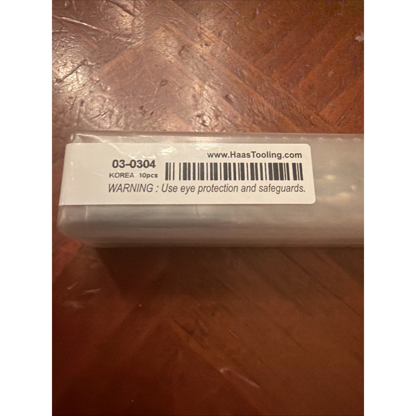 Close-up of a white label on a clear plastic container showing product code 03-0304, origin Korea, quantity 10 pieces, website URL, barcode, and a safety warning to use eye protection and safeguards.