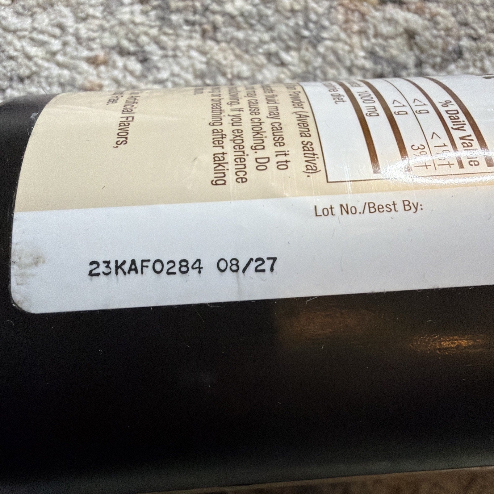 Close-up of a black supplement bottle label showing a lot number "23KAFO284" and a best-by date "08/27" printed on the white part of the label above the bottle surface.