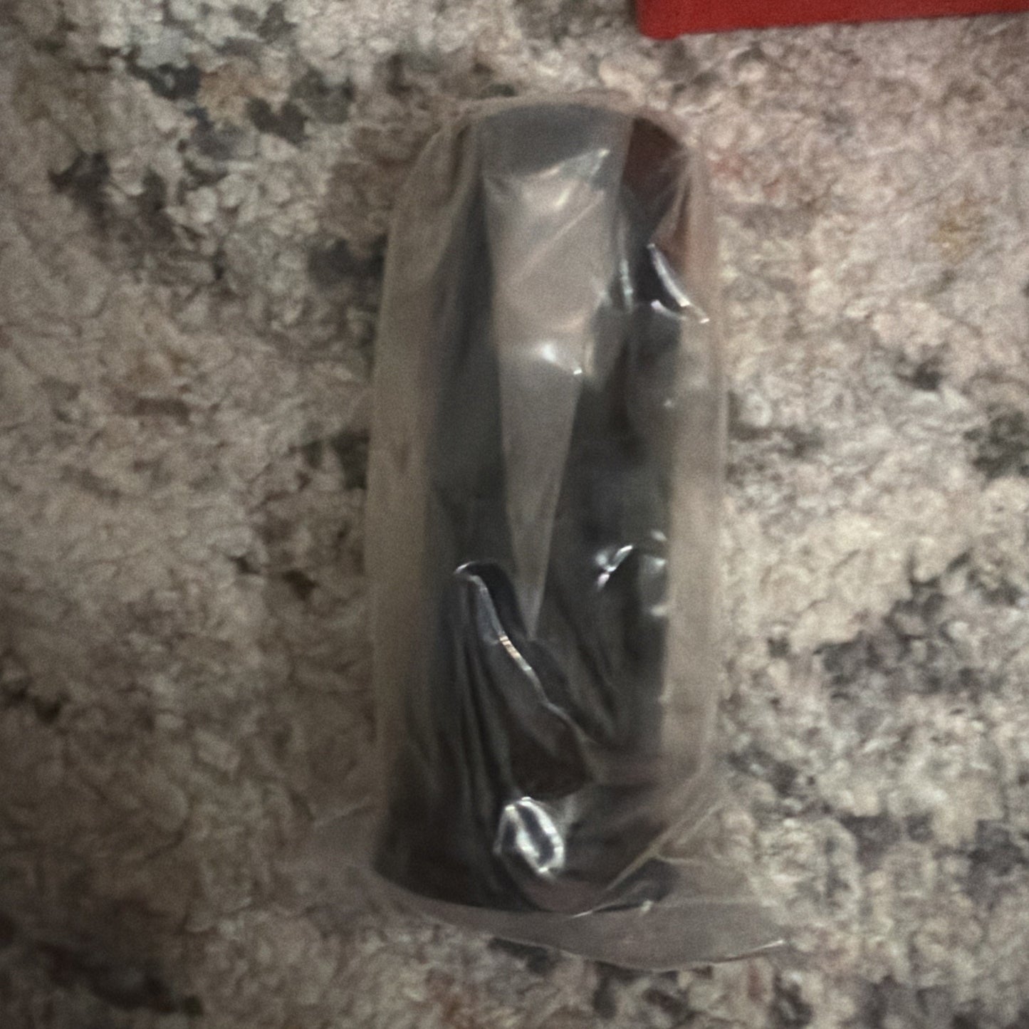 Black cylindrical object wrapped in clear plastic, placed vertically on a light gray and white textured carpet surface.
