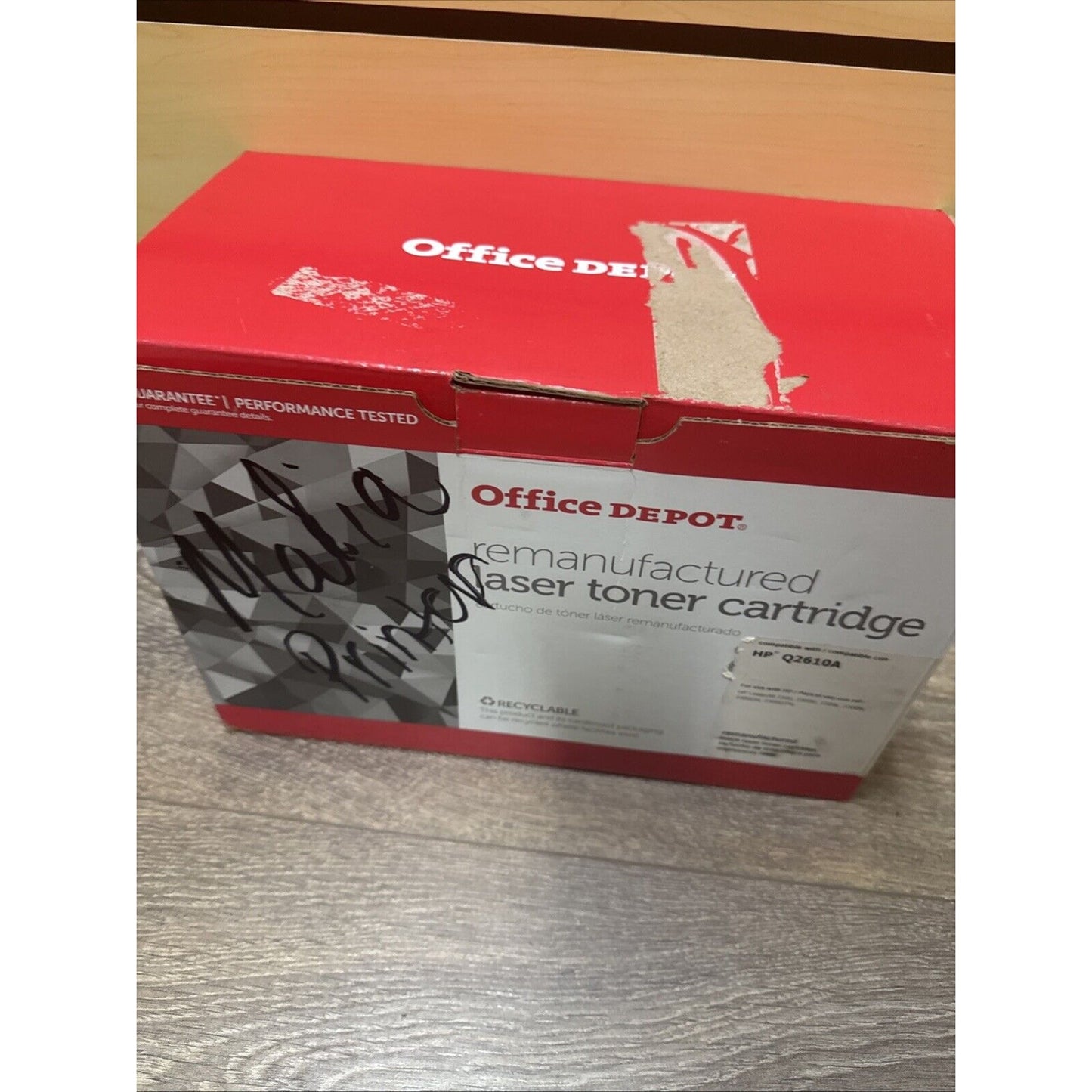 Red and white cardboard box with partially torn label, featuring "Office Depot" branding and text indicating a remanufactured laser toner cartridge, placed on a wooden floor.
