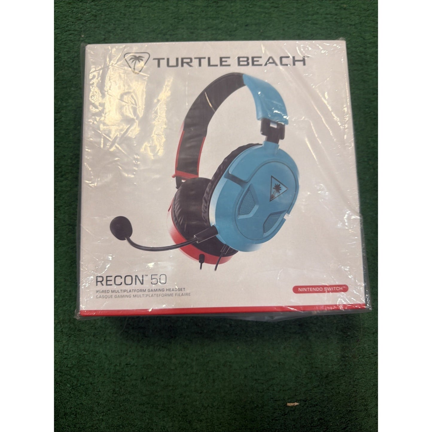 Boxed Turtle Beach Recon 50 wired gaming headset in blue and red, designed for Nintendo Switch, with an adjustable microphone and cushioned ear cups, resting on a green surface.