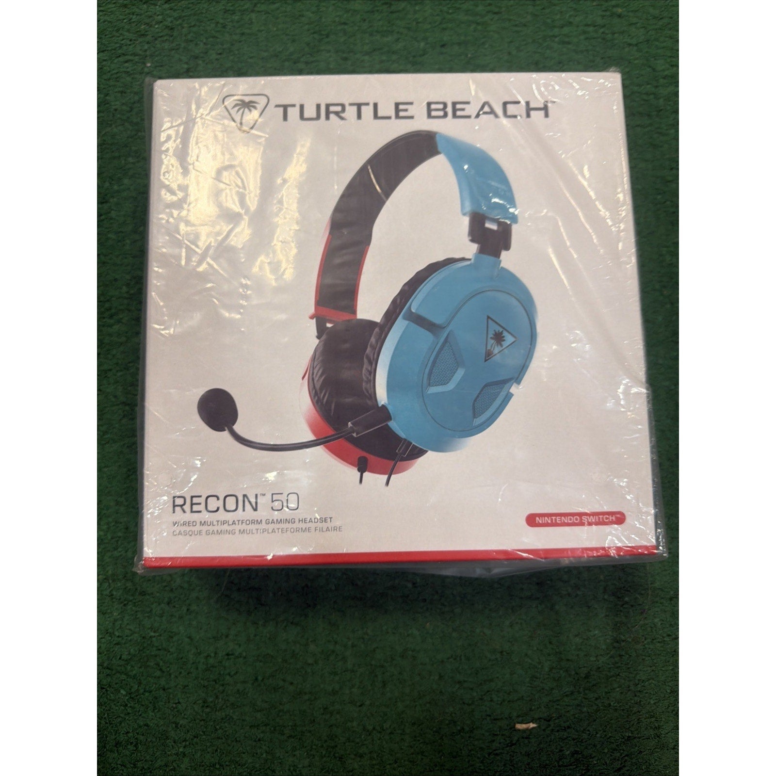 Boxed Turtle Beach Recon 50 wired gaming headset in blue and red, designed for Nintendo Switch, with an adjustable microphone and cushioned ear cups, resting on a green surface.