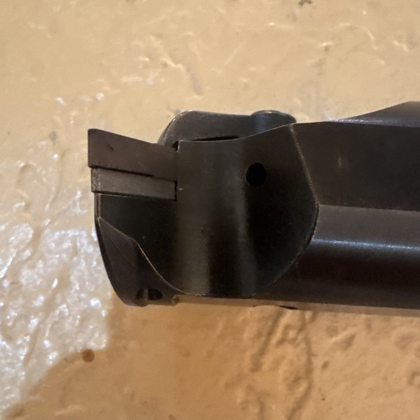 Close-up of a dark metal tool head featuring a small protruding blade or cutting edge, placed against a lightly textured beige background.
