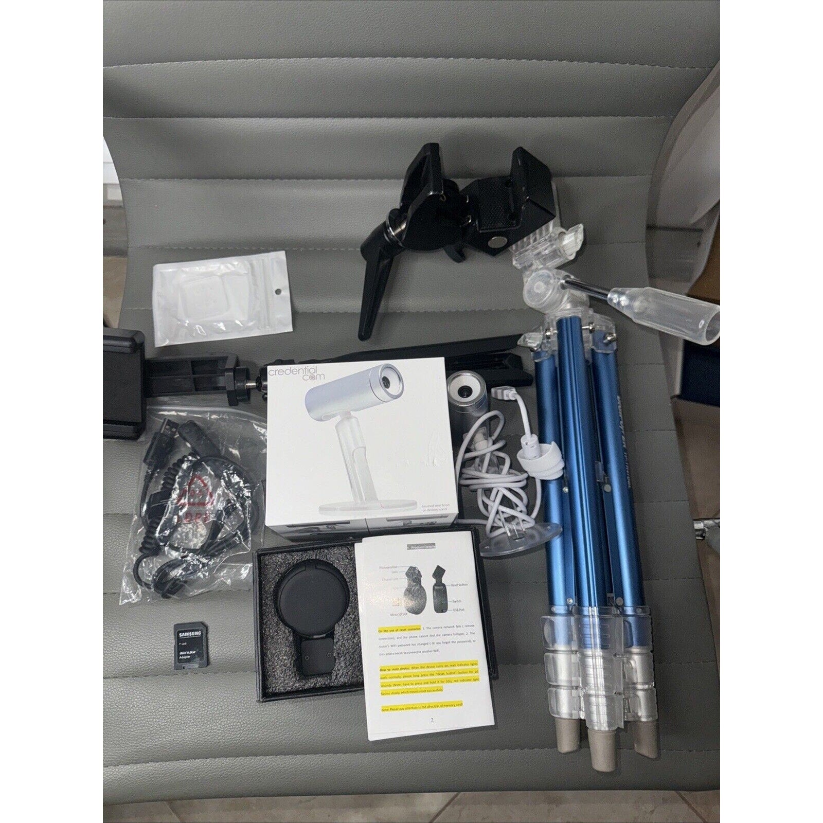 Lot of cameras and tripods including blue tripods, camera accessories, cables, and mounting hardware on a gray chair seat