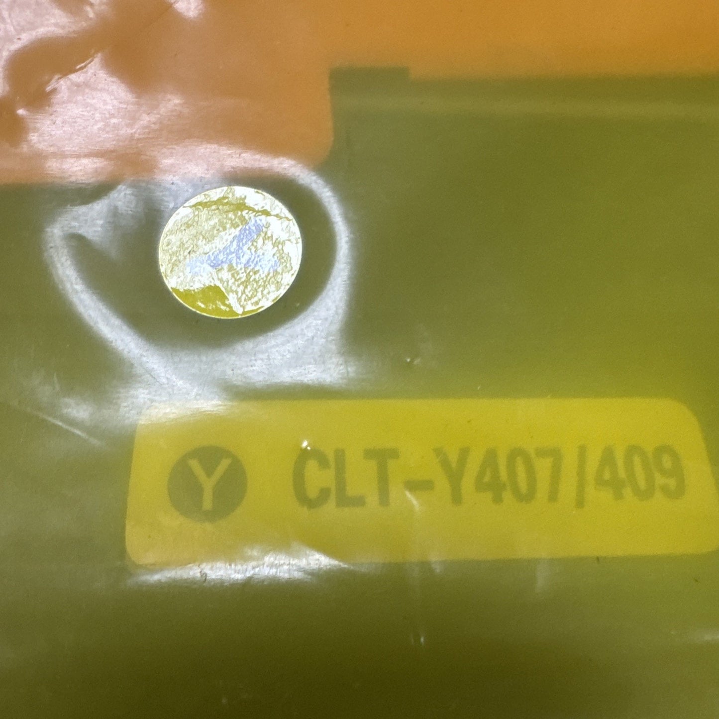 Close-up of a yellow sticker with black text "CLT-Y407/409" and a black "Y" inside a circle, placed on a shiny surface near a small round reflective area.