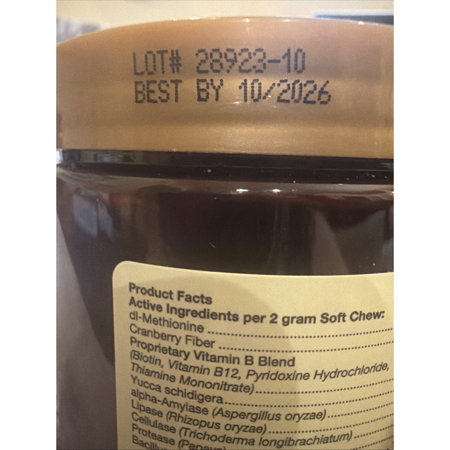 Close-up of a brown jar with a gold lid showing a printed lot number and best-by date, along with a product label listing active ingredients including vitamins, enzymes, and cranberry fiber for soft chews.