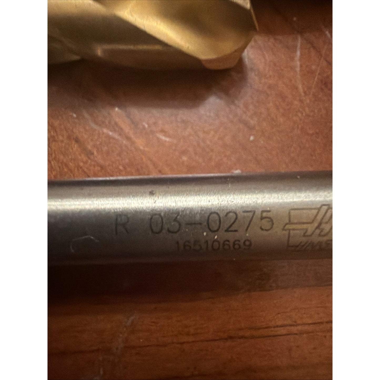 Close-up of a metal tool shaft engraved with the code "R 03-0275" and a serial number, resting on a wooden surface with part of a gold-colored drill bit visible in the background.