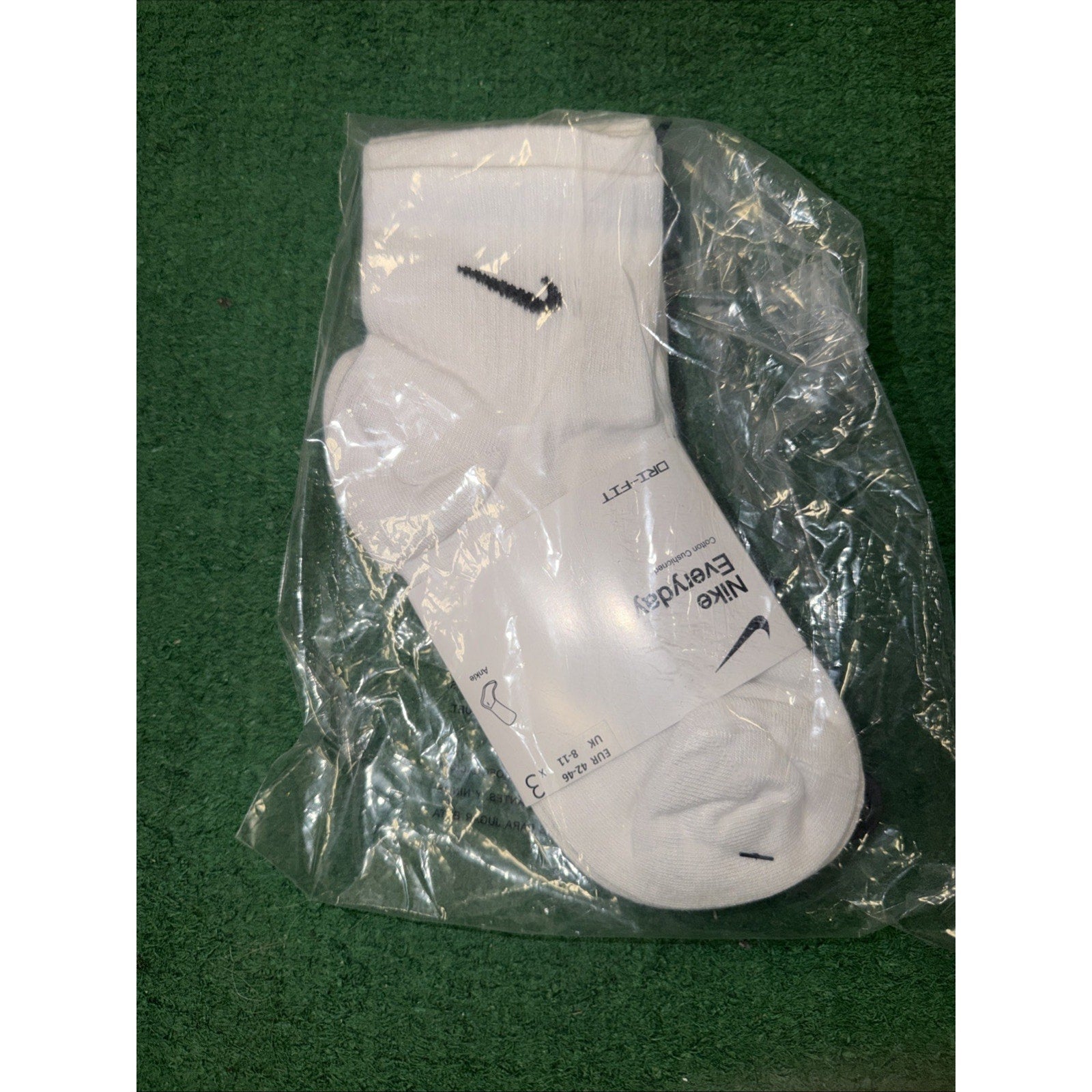 Pair of white Nike Everyday cushioned ankle socks with black swoosh logos on a green textured surface, still sealed in clear plastic packaging.