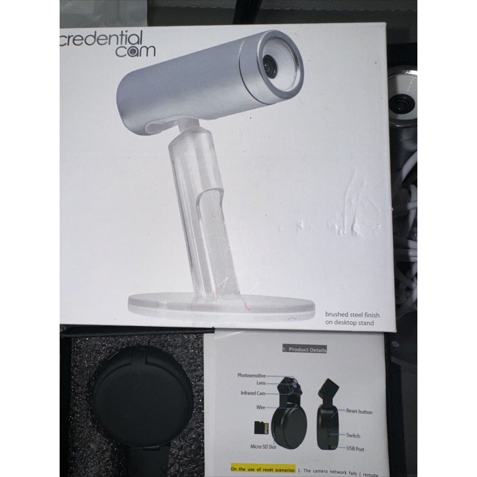 Silver cylindrical Credential Cam security camera with clear desktop stand and product details card in foam packaging