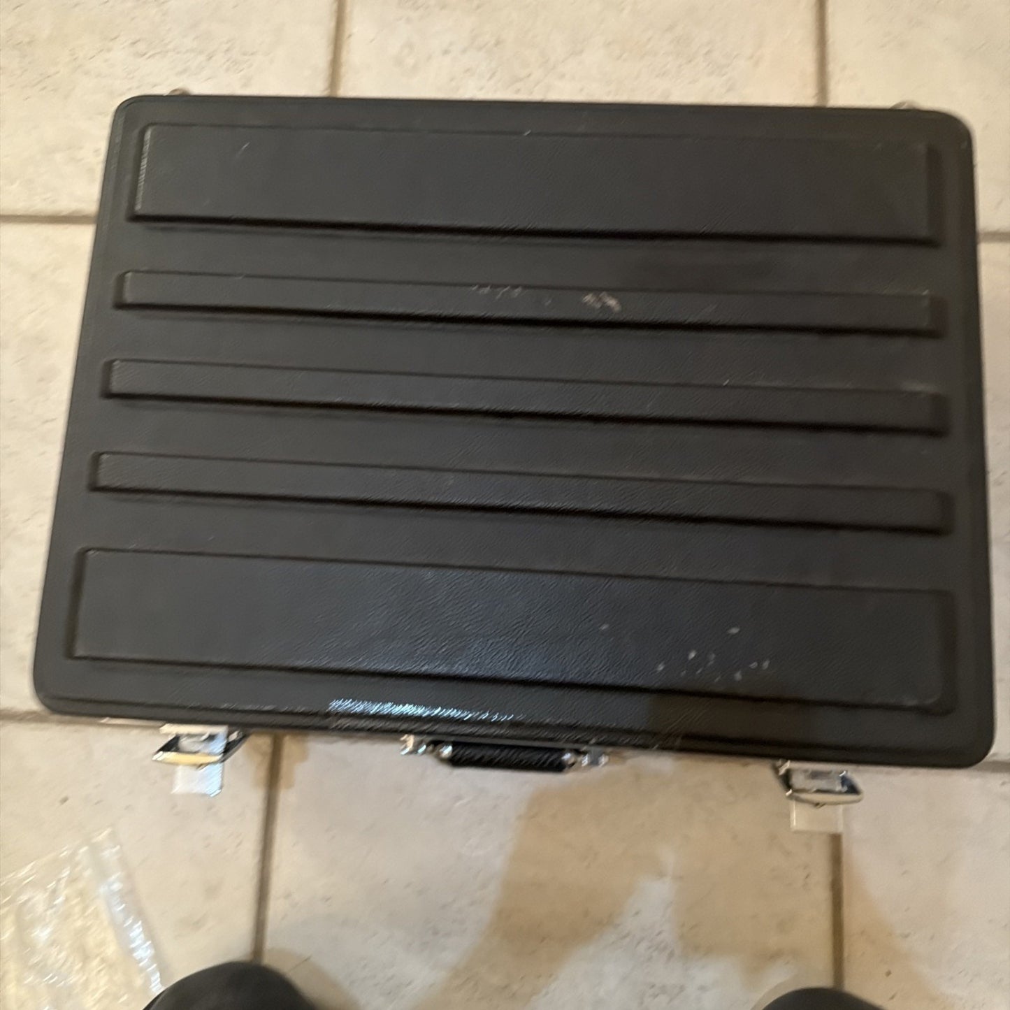 Black hard plastic carrying case with horizontal raised ridges and two silver metal latches, placed on a tiled floor.