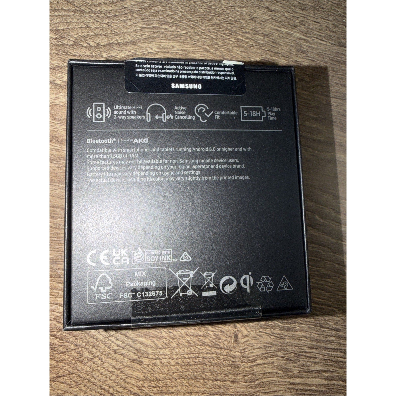 Black Samsung product box back featuring icons for Hi-Fi 2-way speakers, active noise cancelling, Bluetooth by AKG compatibility, battery life of 5 to 18 hours, and multiple certification logos on a wood surface.