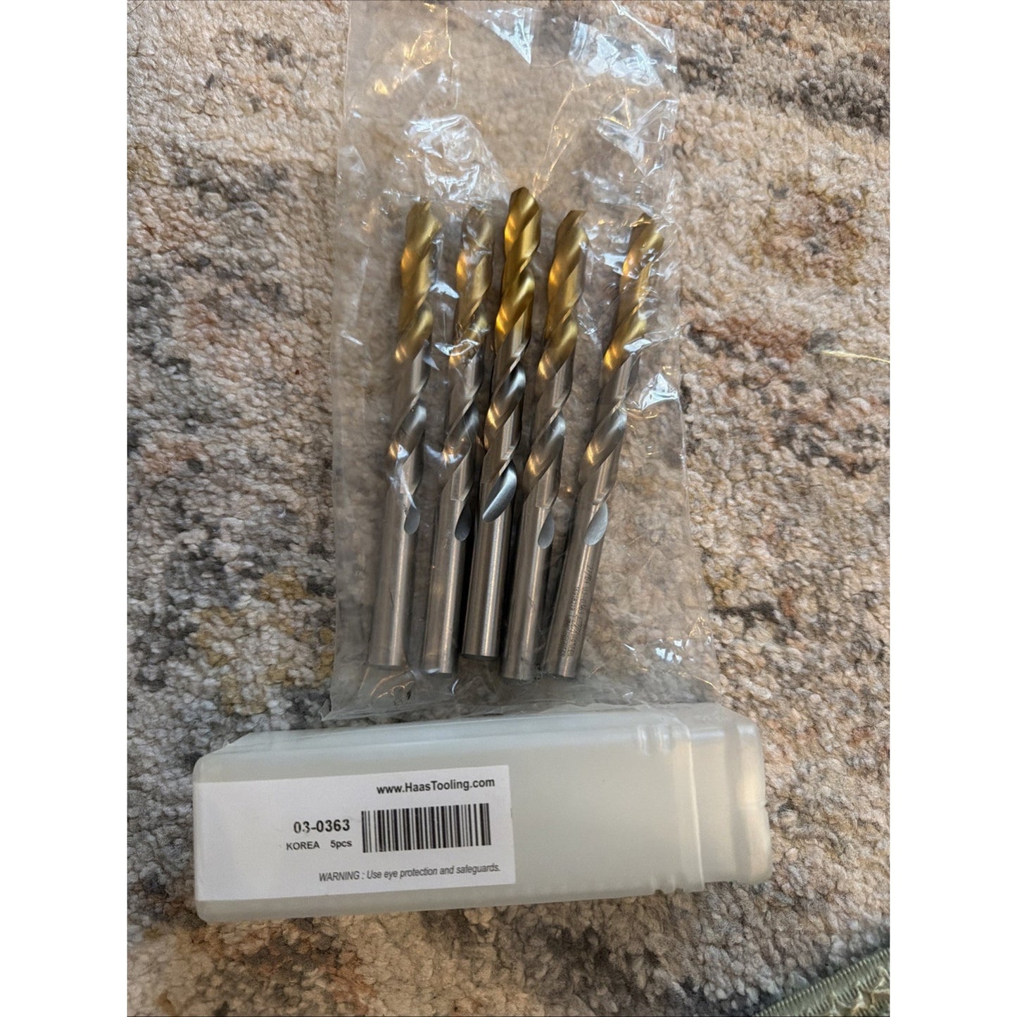 Set of eight drill bits with gold-colored coated tips and shiny silver shanks, arranged upright inside a clear plastic bag, placed on a textured carpet next to a white plastic storage case.