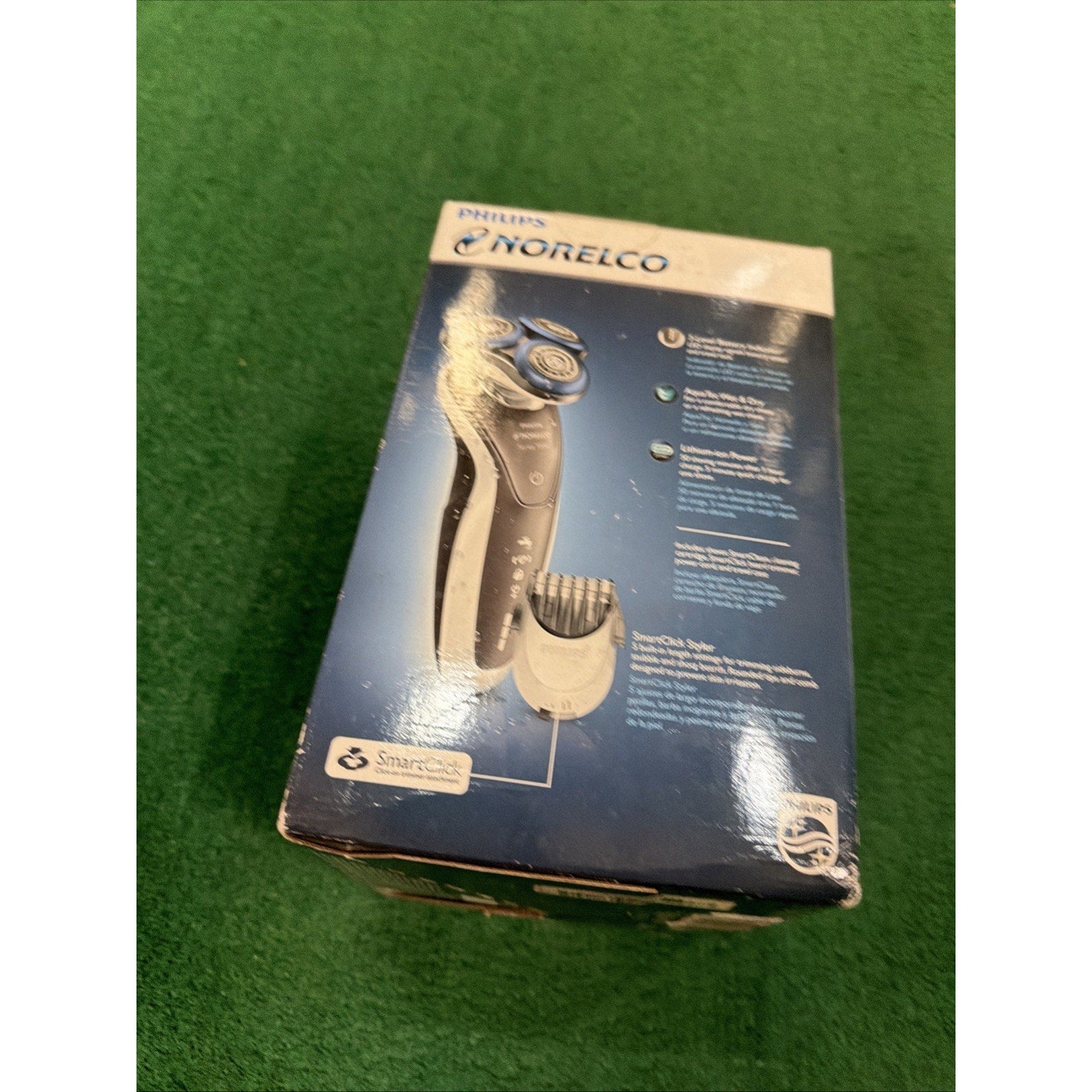 Box packaging of a Philips Norelco electric shaver featuring an image of the shaver and trimming attachment against a blue background, placed on a green surface.
