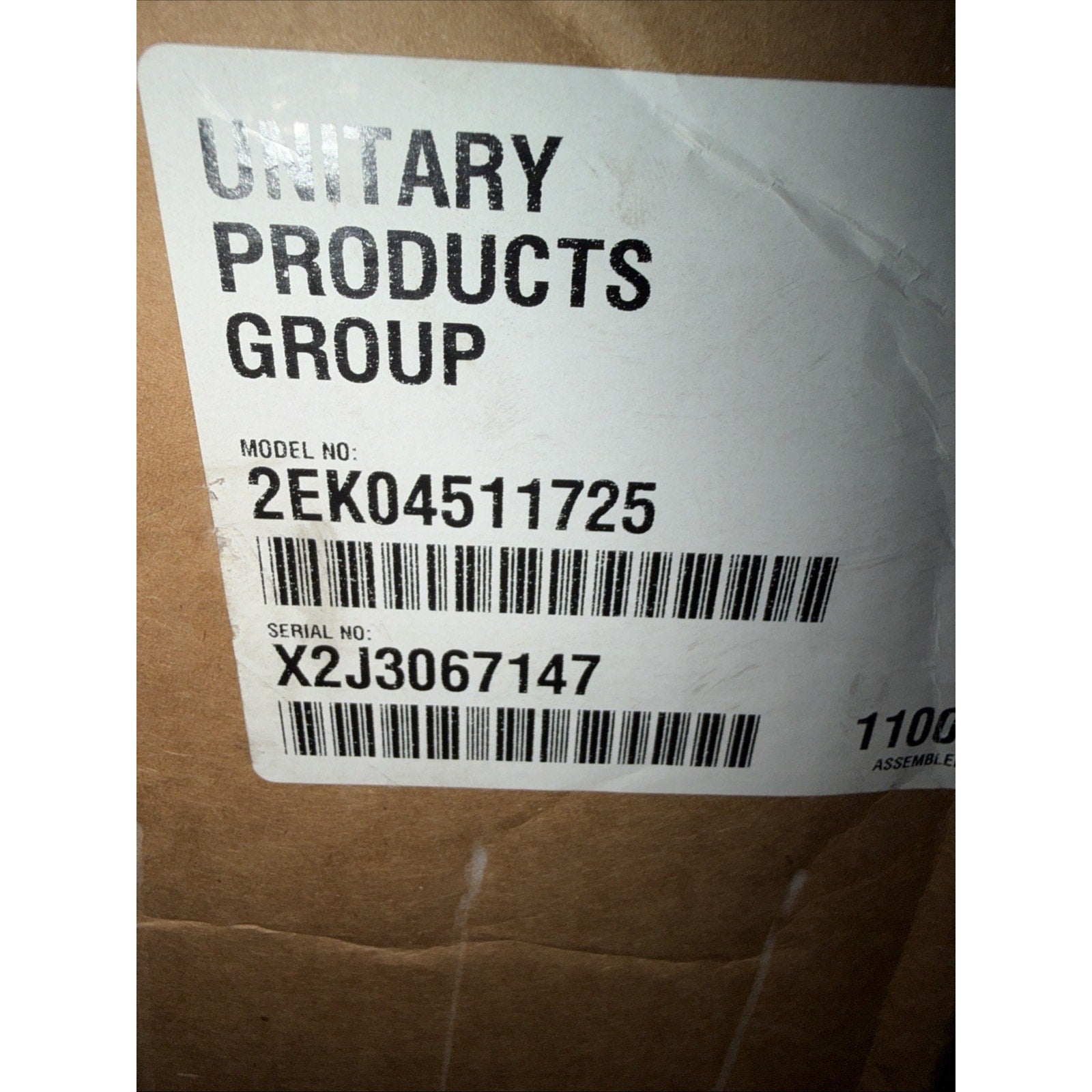 Close-up view of a white label on a brown cardboard box showing the brand name Unitary Products Group with printed model number 2EK04511725 and serial number X2J3067147 alongside two barcodes.