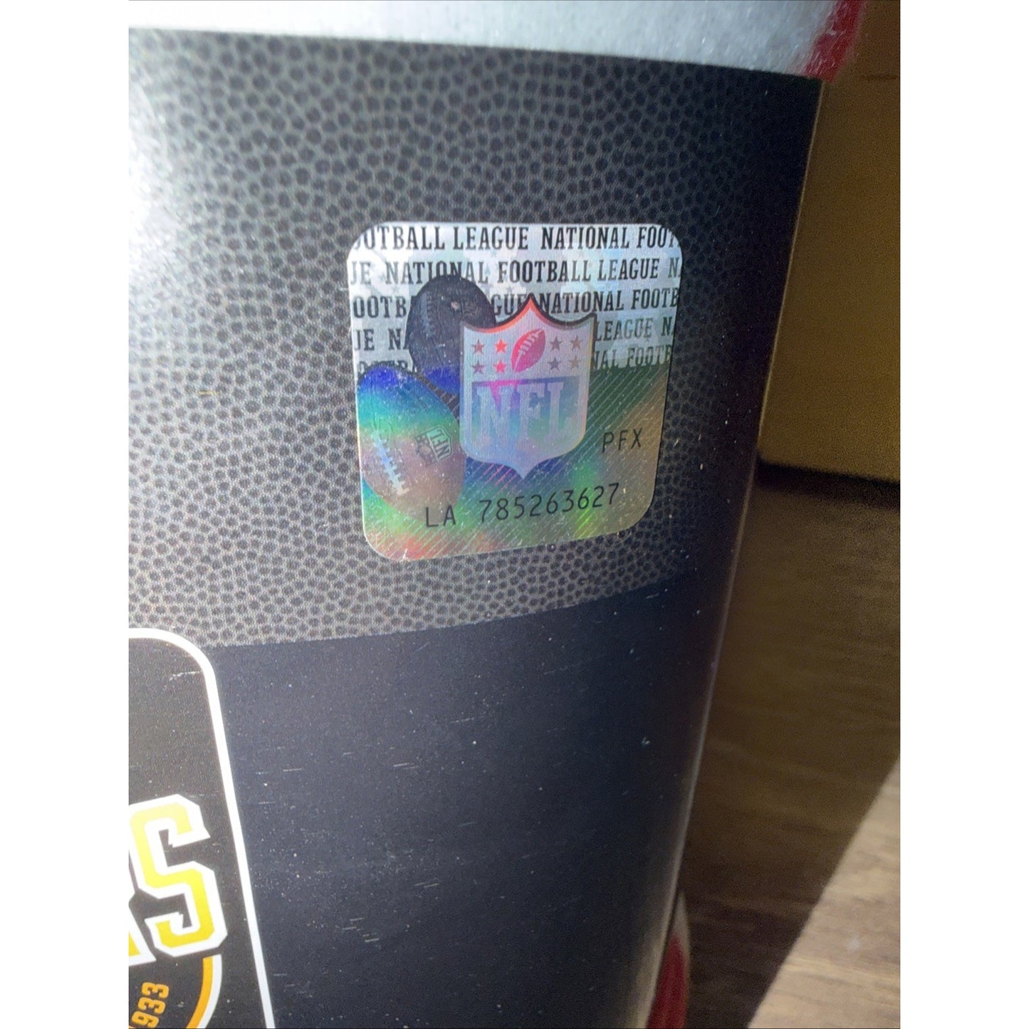 Close-up of an NFL holographic authenticity sticker with a serial number on a textured black and gray surface, illuminated by natural light.