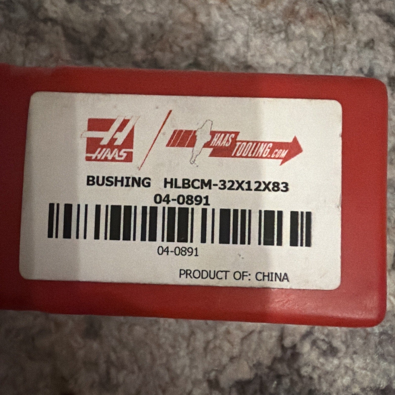 White label with black text and barcodes adhered to a red rectangular plastic container, showing product code information and origin marked as China, on a gray textured surface.