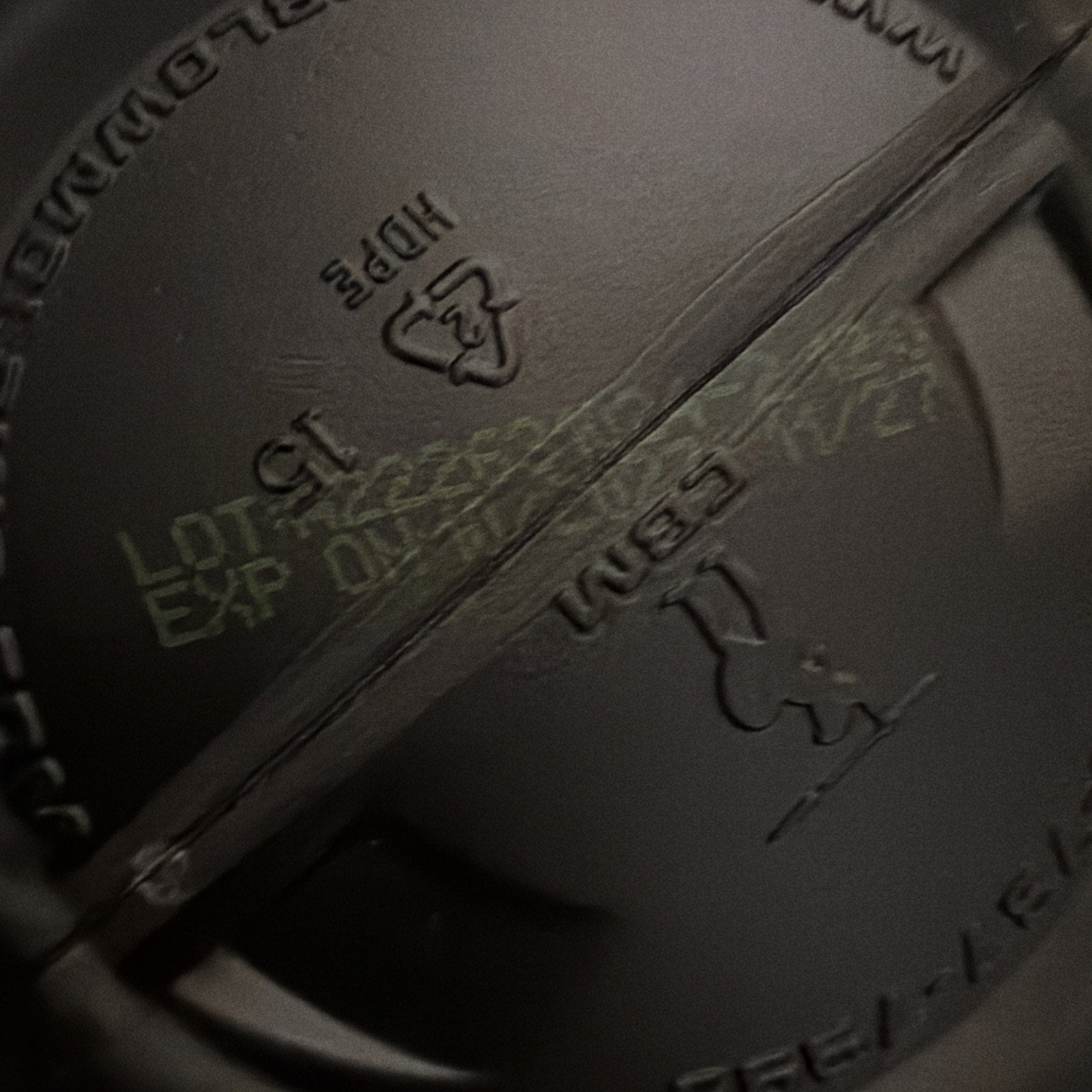 Close-up of pale yellow batch code and expiration date printed on the inside bottom of a dark plastic container lid with molded recycling and material symbols.