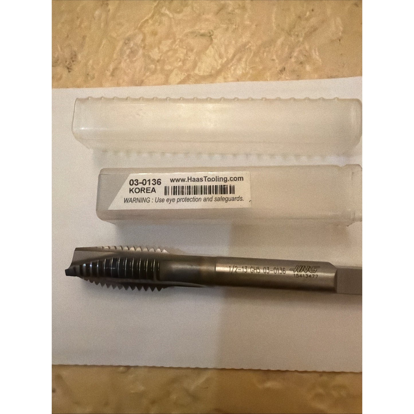 Metal threading tap with a square drive shank and sharp-cut threads, displayed below a clear plastic storage case labeled with product code, website, and safety warning on a light surface.