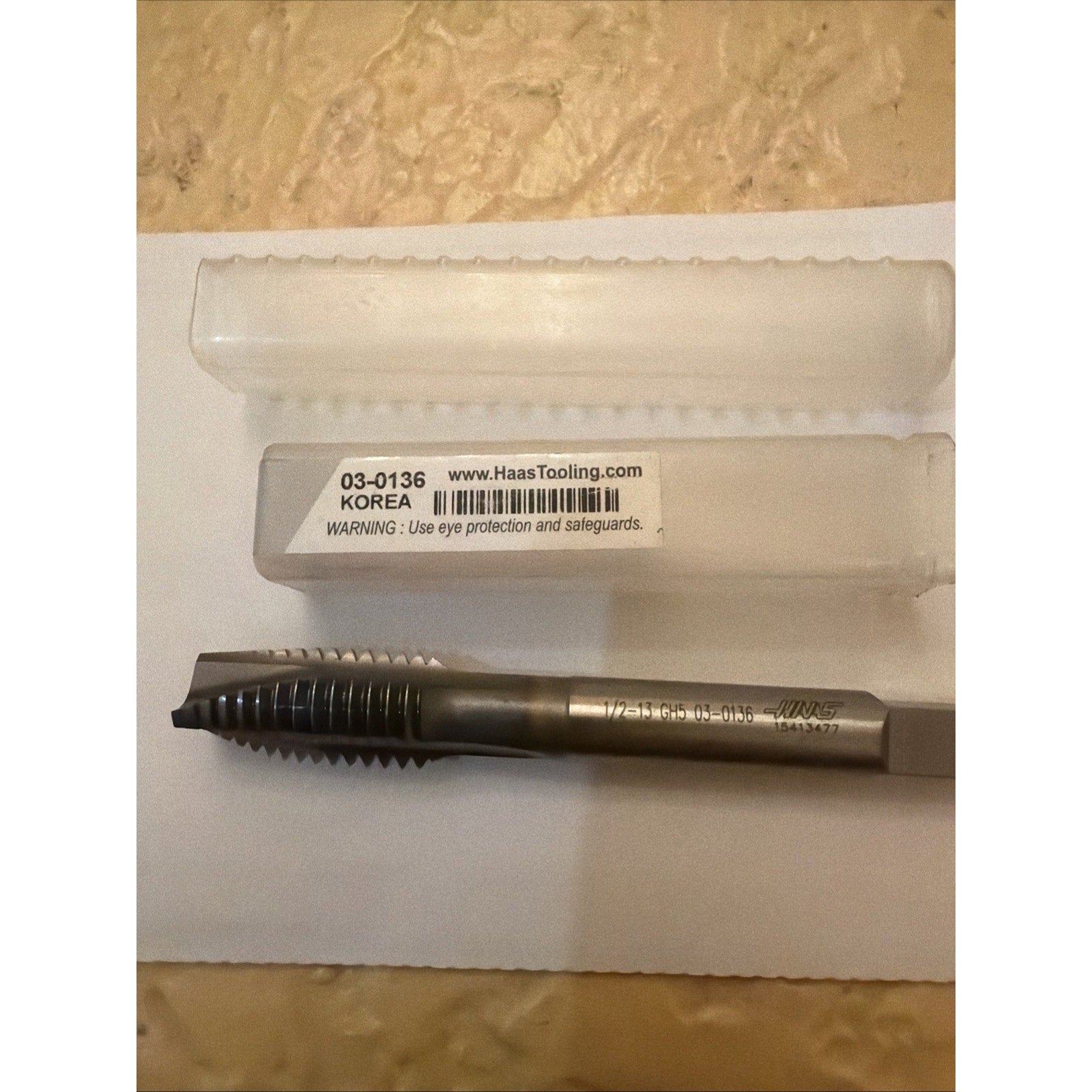 Metal threading tap with a square drive shank and sharp-cut threads, displayed below a clear plastic storage case labeled with product code, website, and safety warning on a light surface.