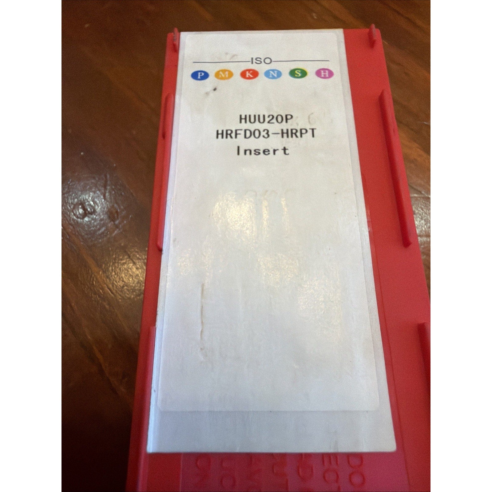 Red plastic storage case with a white label displaying ISO classification symbols in multiple colors and text "HUU20P HRFD03-HRPT Insert," placed on a wooden surface.