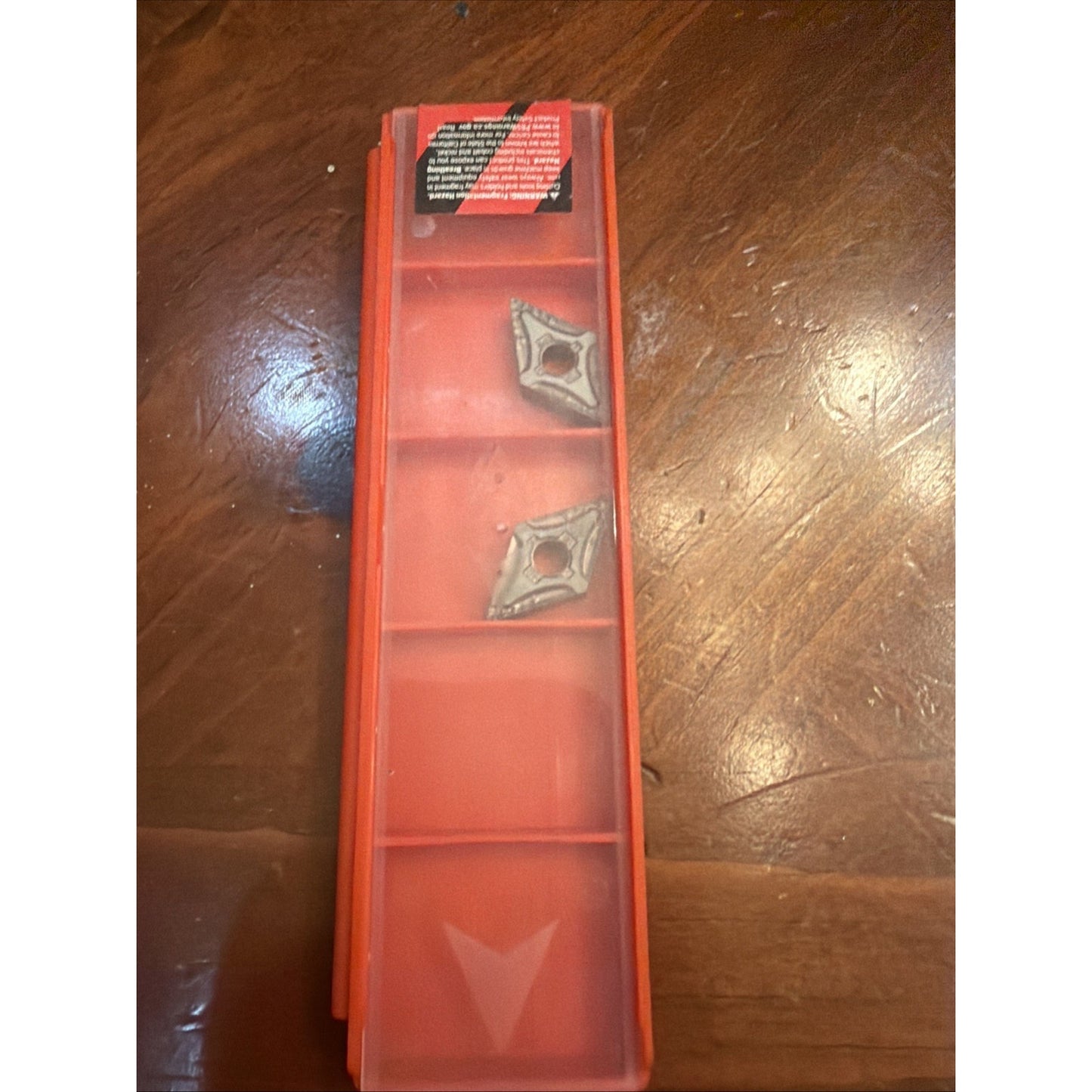 Long translucent red plastic storage case with two triangular metal cutting inserts placed in separate compartments, resting on a wooden surface with visible grain.