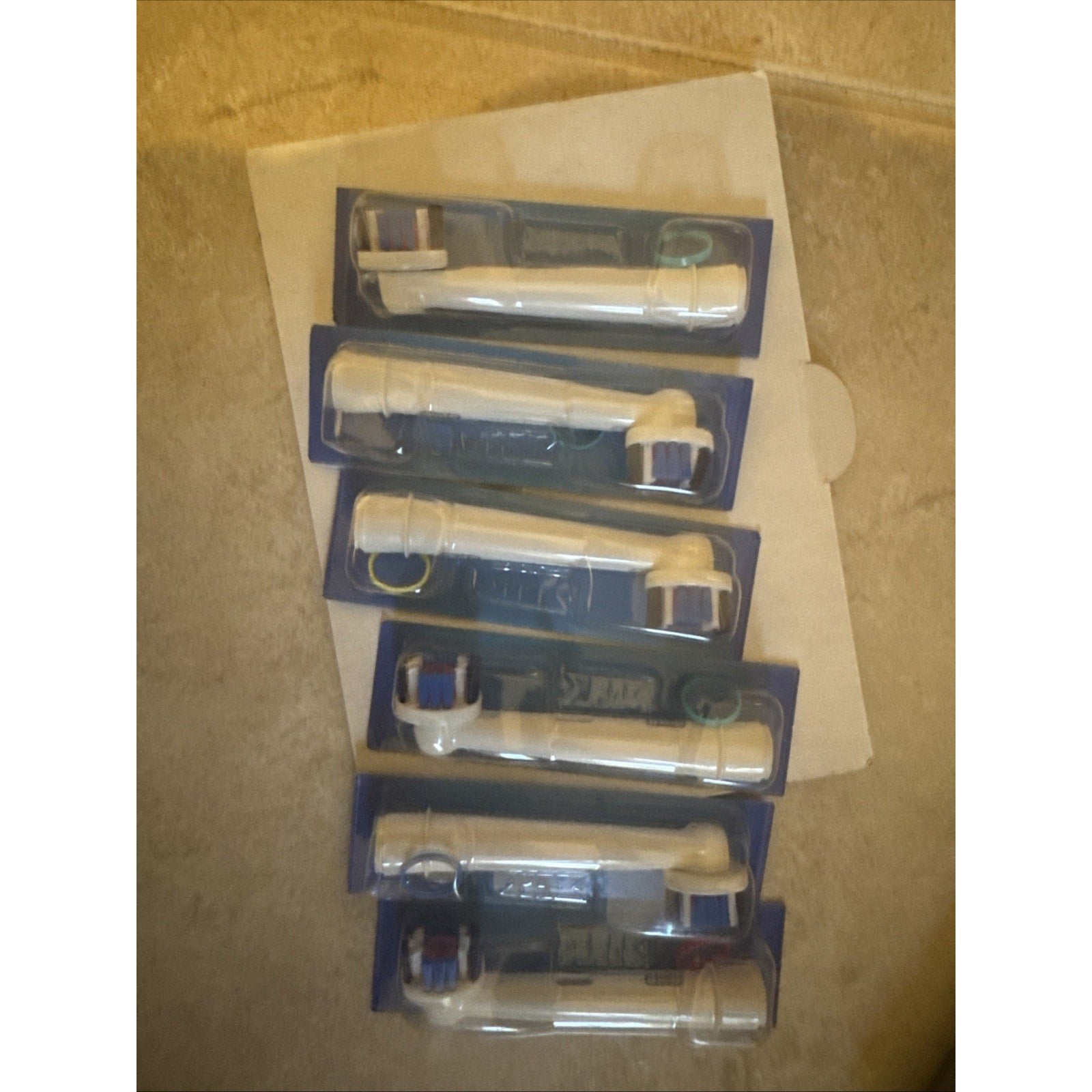 Six Oral-B replacement electric toothbrush heads in plastic packaging arranged on a beige surface
