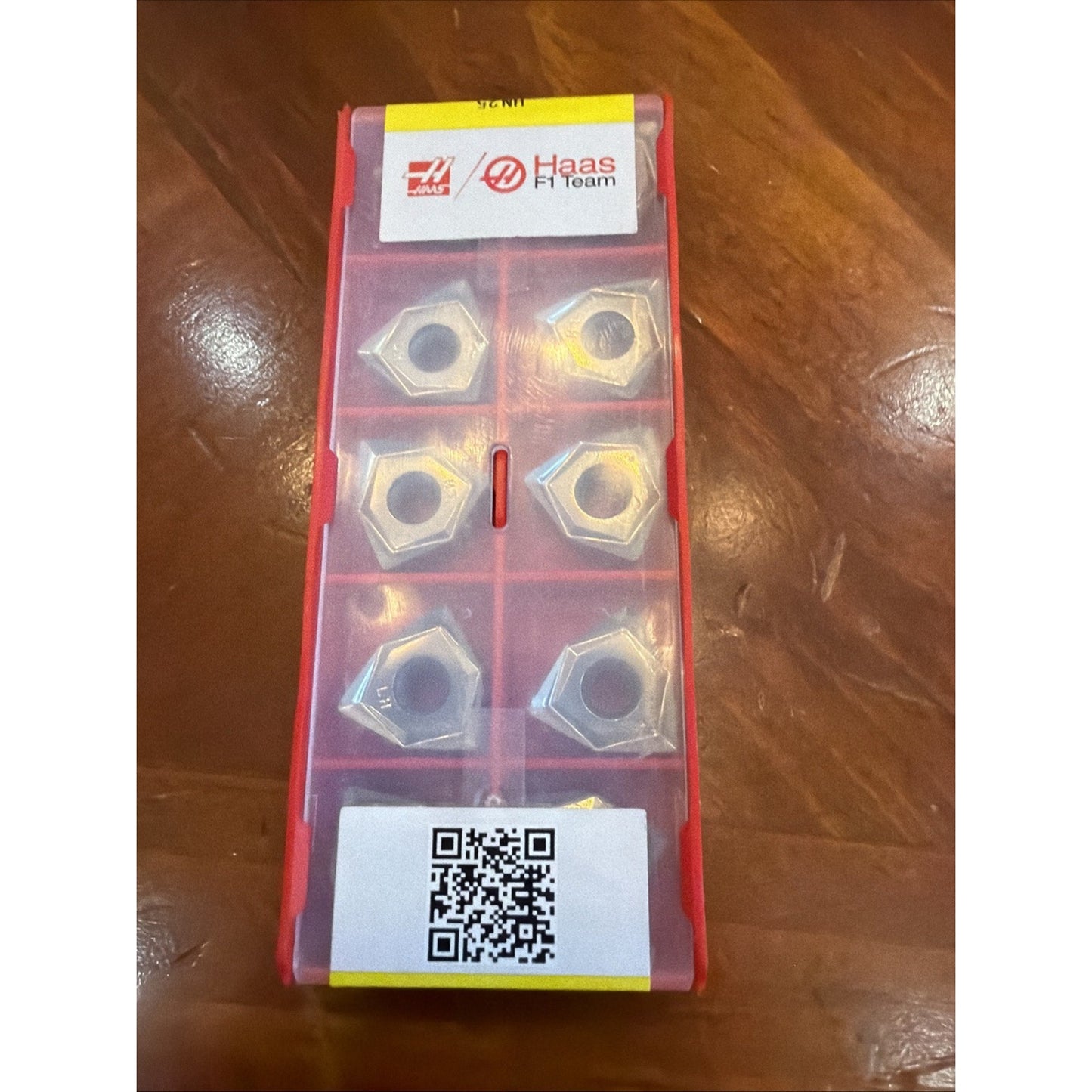 Red plastic container holding six silver metal nuts, with a Haas F1 Team label and a QR code sticker on a wooden surface.