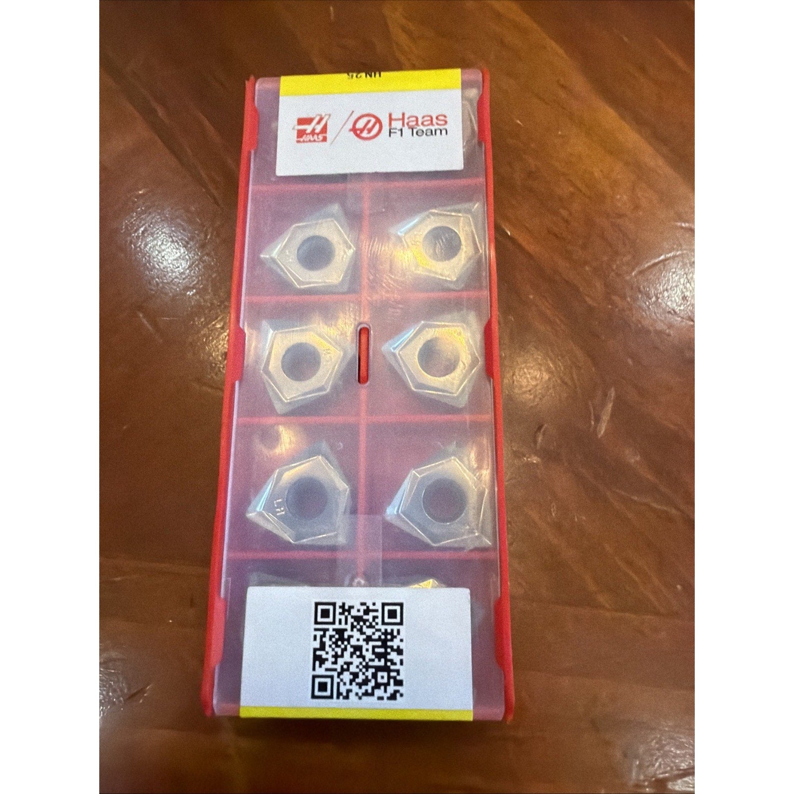 Red plastic container holding six silver metal nuts, with a Haas F1 Team label and a QR code sticker on a wooden surface.