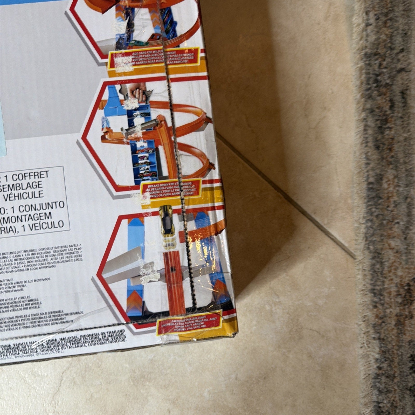Corner of a toy track box displaying images of orange and blue race tracks with cars, featuring looped sections and a vertical launch ramp on a tiled floor next to a carpet edge.