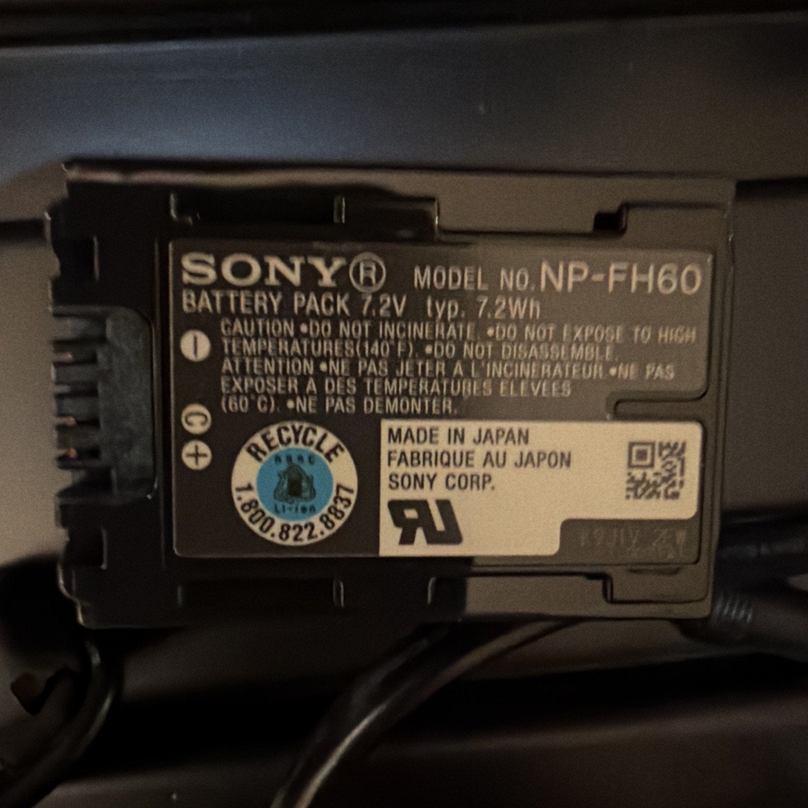 Close-up of a Sony rechargeable lithium-ion battery pack labeled NP-FH60 with safety warnings, certification marks, and "Made in Japan" text on a black device.