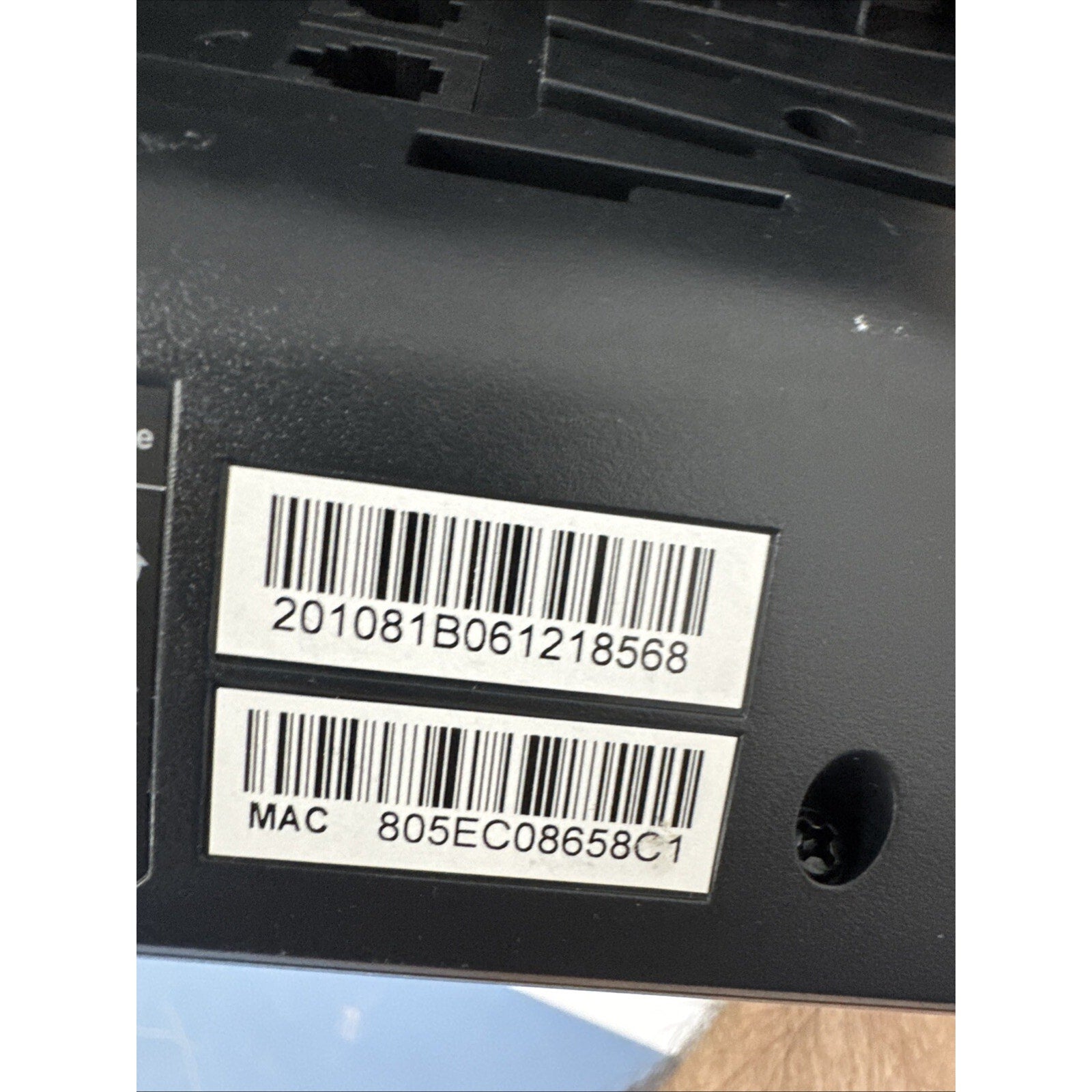 Close-up view of two white barcode labels with black alphanumeric codes on a matte black plastic surface, alongside a small screw hole.
