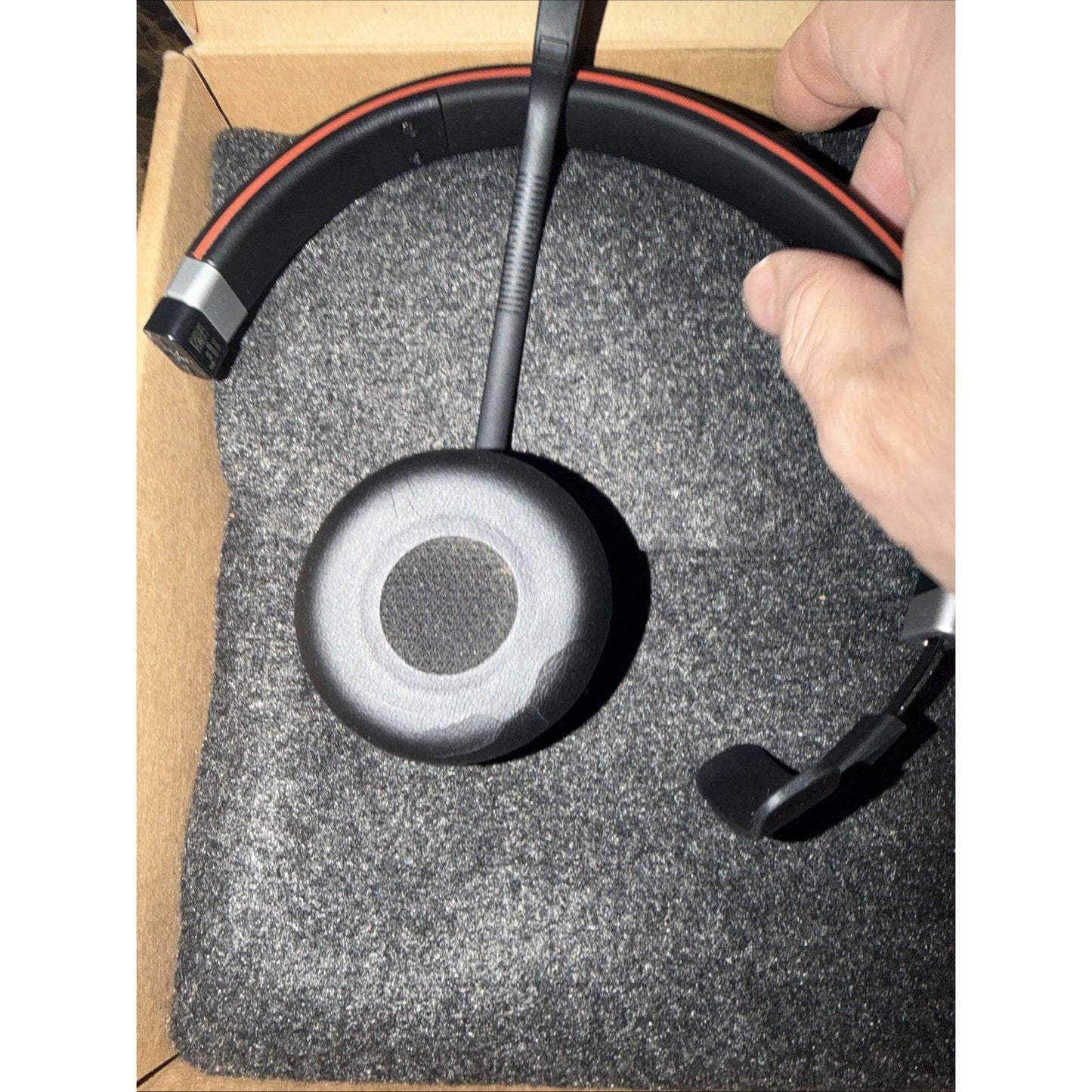 Black over-the-head headset with a microphone boom, cushioned ear cup, and red accent on the headband, resting on gray foam inside a brown cardboard box.