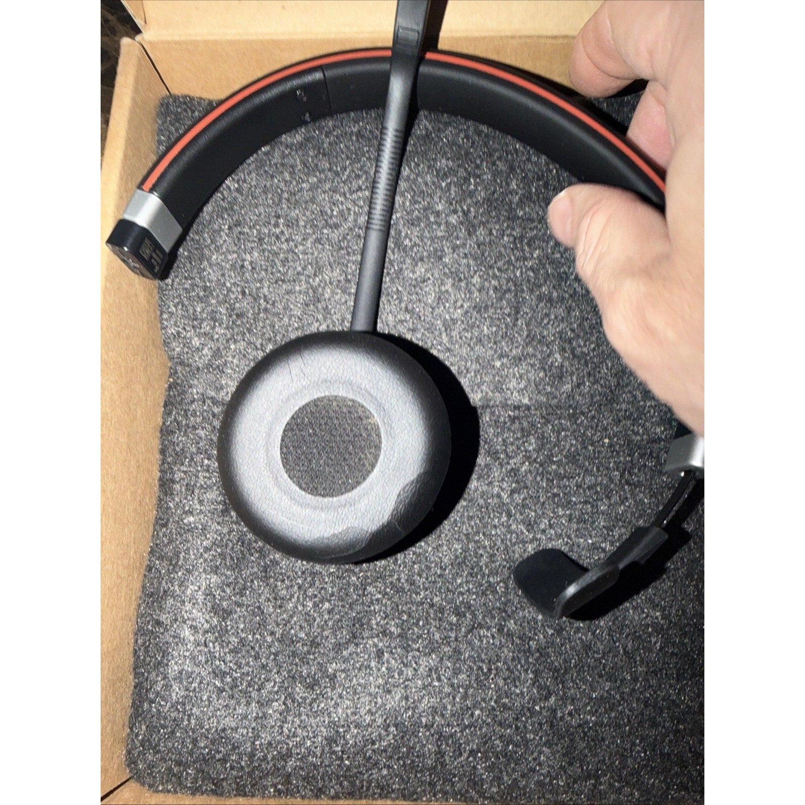 Black over-the-head headset with a microphone boom, cushioned ear cup, and red accent on the headband, resting on gray foam inside a brown cardboard box.