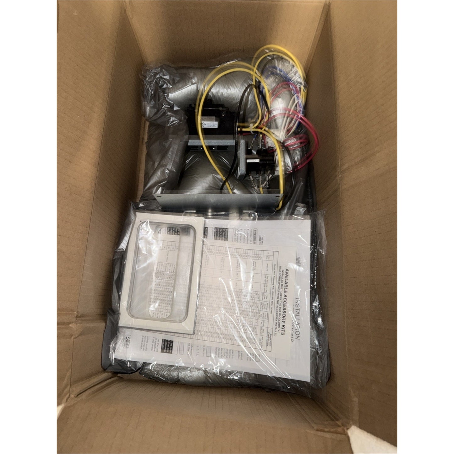 Top view of an open cardboard box containing a packaged mechanical or electrical component with connected wires, wrapped in protective plastic and accompanied by printed instruction sheets.