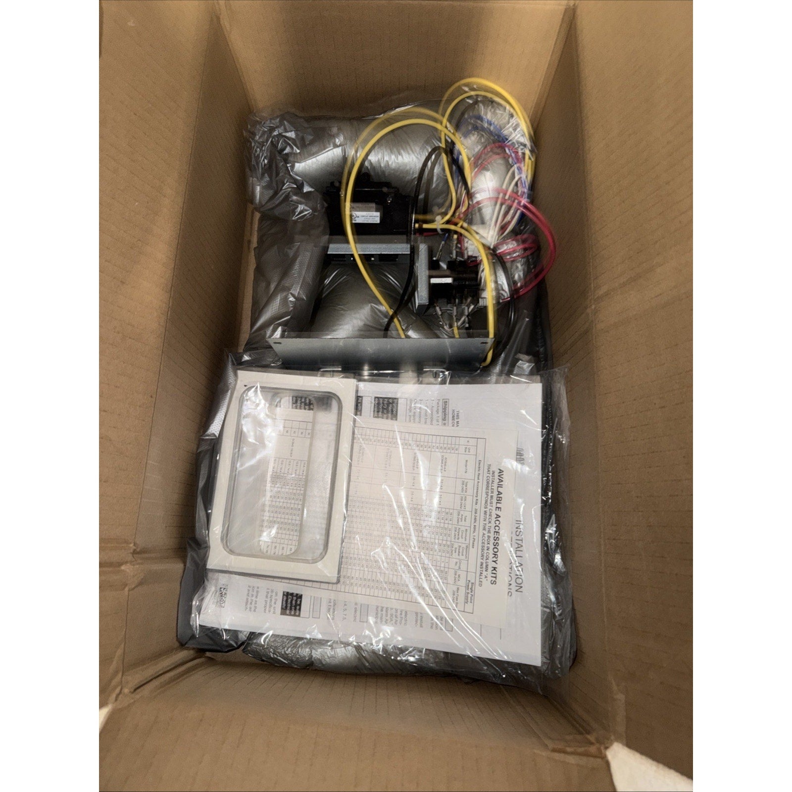 Top view of an open cardboard box containing a packaged mechanical or electrical component with connected wires, wrapped in protective plastic and accompanied by printed instruction sheets.