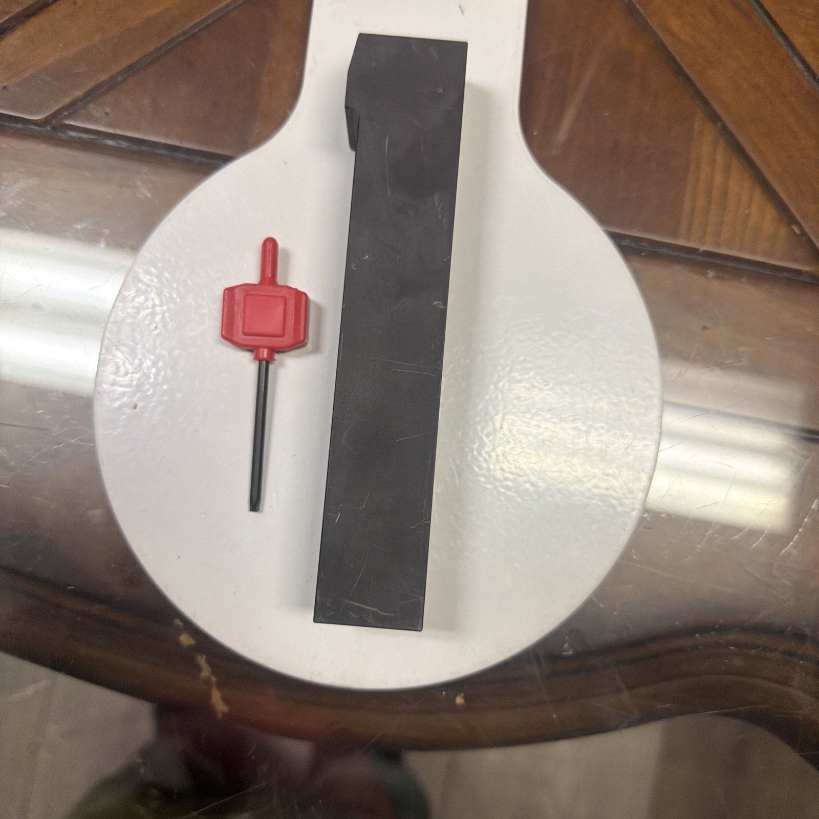 Black rectangular metal tool with a flat top surface, placed next to a small red plastic-handled hex wrench on a circular white background over a glass tabletop.