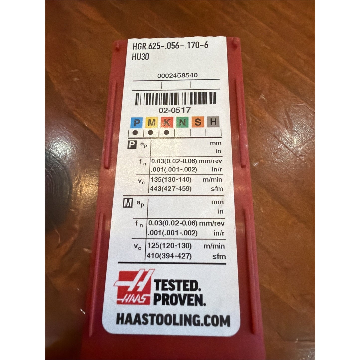 Red plastic case with a white label showing product code "HGR.625-.056-.170-6 HU30," machining parameters for different materials, colored ISO classification squares, barcode, Haas Tested Proven logo, and website on a wooden surface.