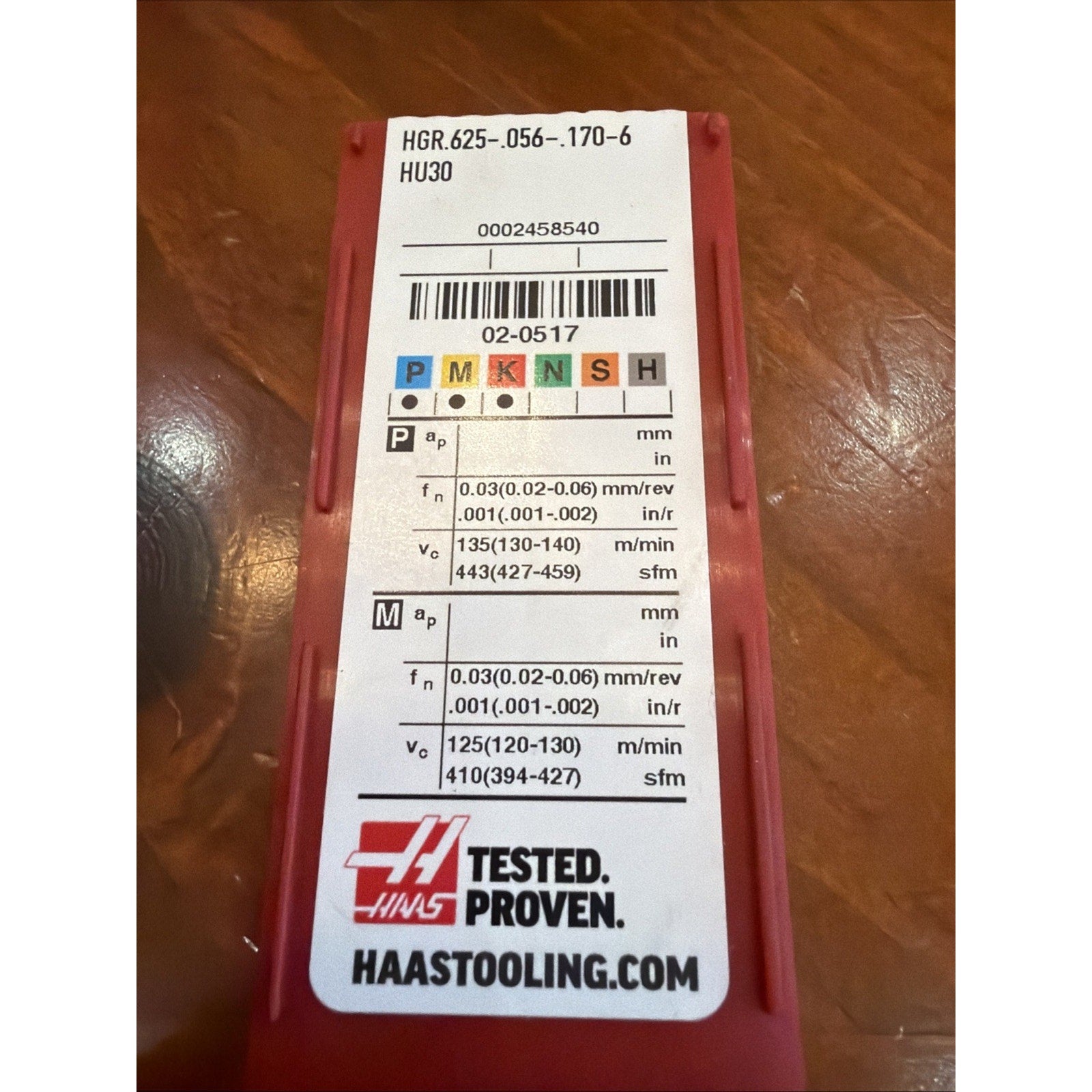 Red plastic case with a white label showing product code "HGR.625-.056-.170-6 HU30," machining parameters for different materials, colored ISO classification squares, barcode, Haas Tested Proven logo, and website on a wooden surface.