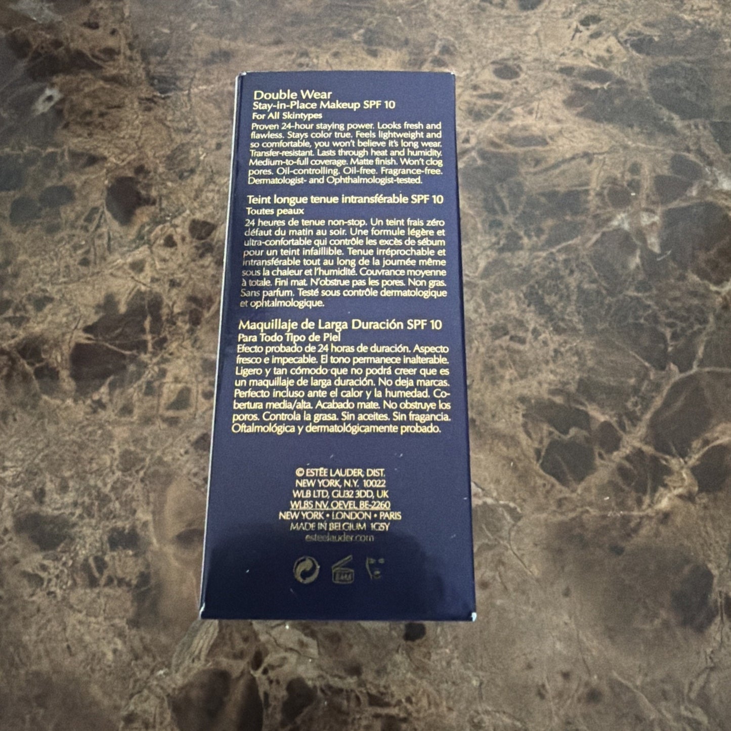 Dark blue rectangular box with detailed product information in English, French, and Spanish describing a 24-hour wear, SPF 10 stay-in-place makeup, placed on a marble surface.