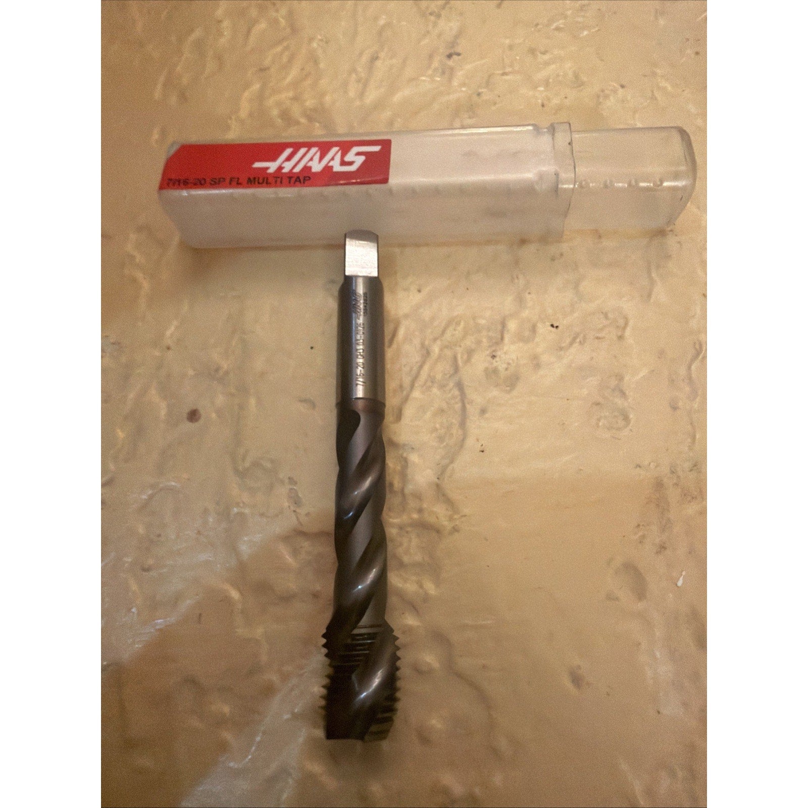 Dark metal spiral threading tap with a square drive end, positioned vertically beneath a clear plastic storage case labeled with a red and white Haas logo on a textured beige surface.