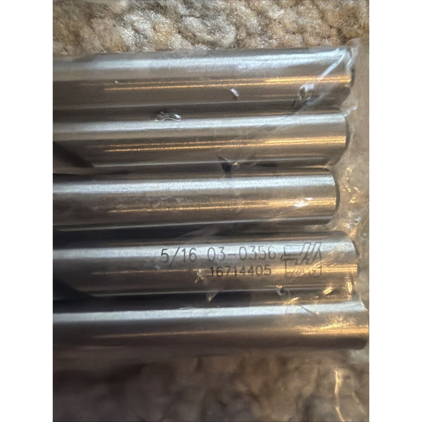 Close-up of five metallic drill or cutting tool bars laid horizontally on a soft textured surface, with visible engraved numbers and letters on one bar, encased partially in clear plastic.