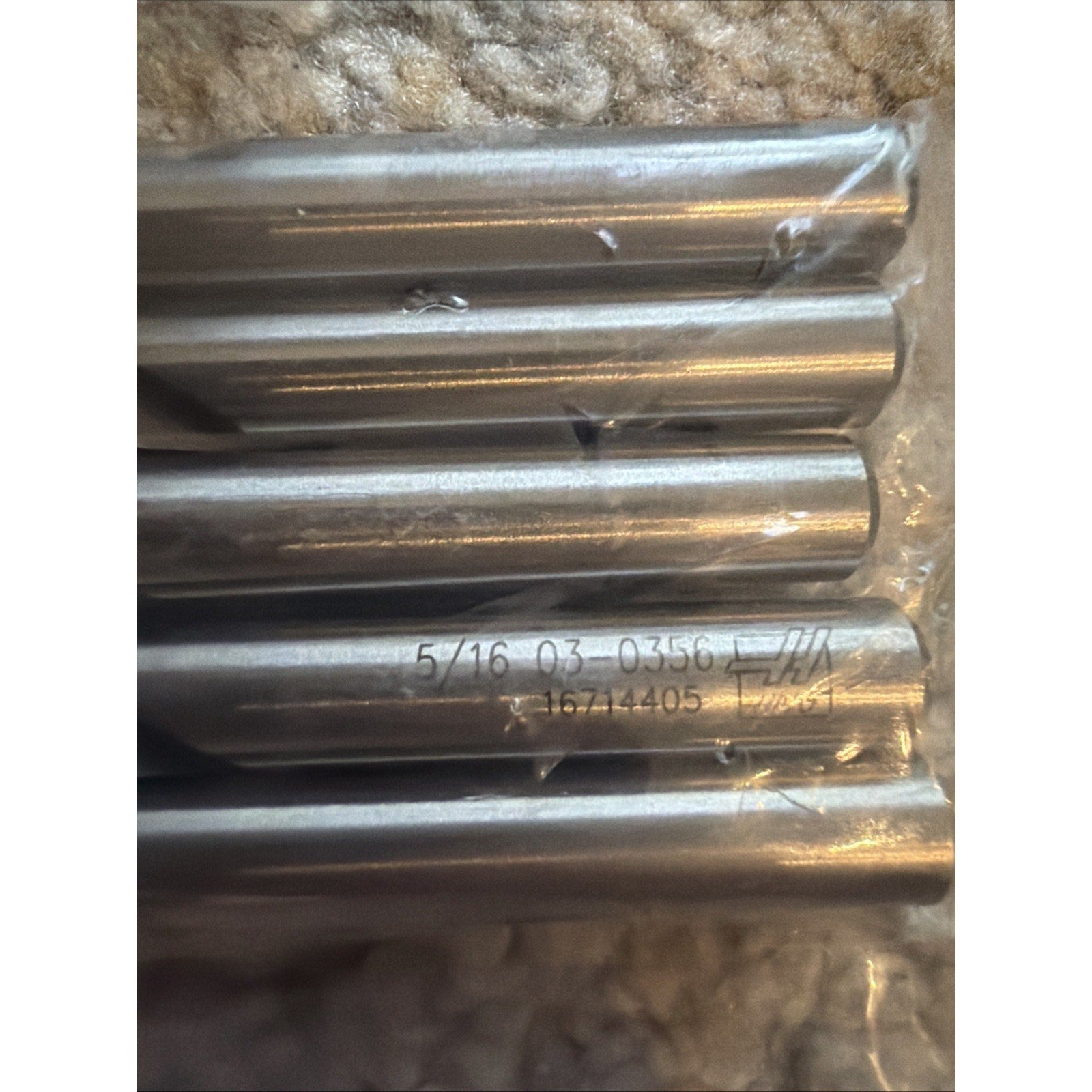 Close-up of five metallic drill or cutting tool bars laid horizontally on a soft textured surface, with visible engraved numbers and letters on one bar, encased partially in clear plastic.