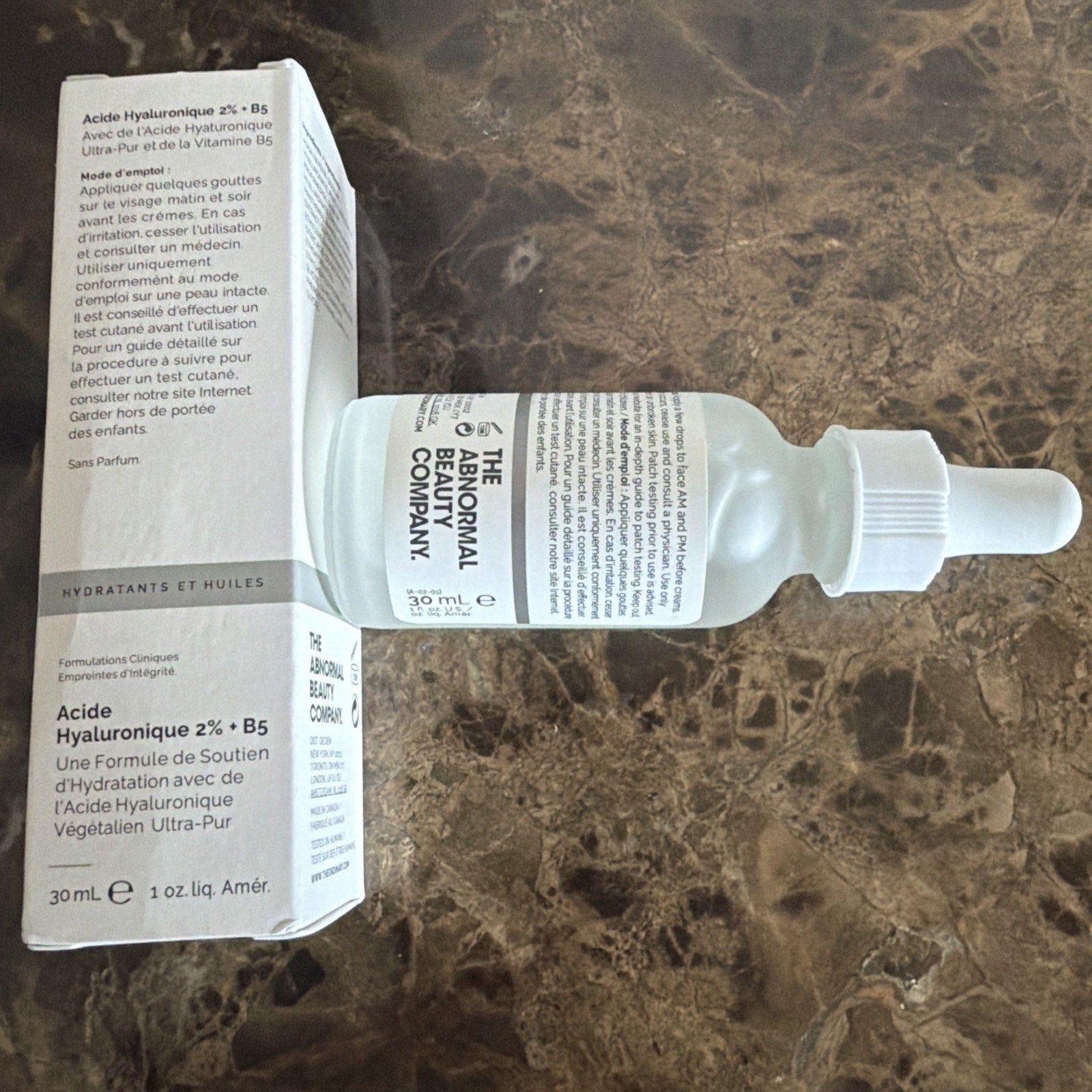 Transparent frosted glass dropper bottle with white cap containing hyaluronic acid serum, placed on a brown marble surface next to a white rectangular box with product information in French and English.